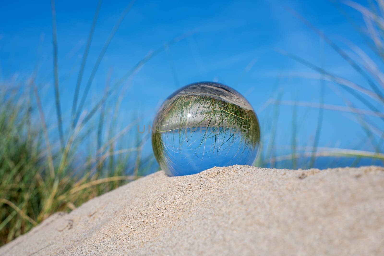 A glass ball lies in the sand of a dune by the sea, the dune grass and the blue sky are reflected in the ball