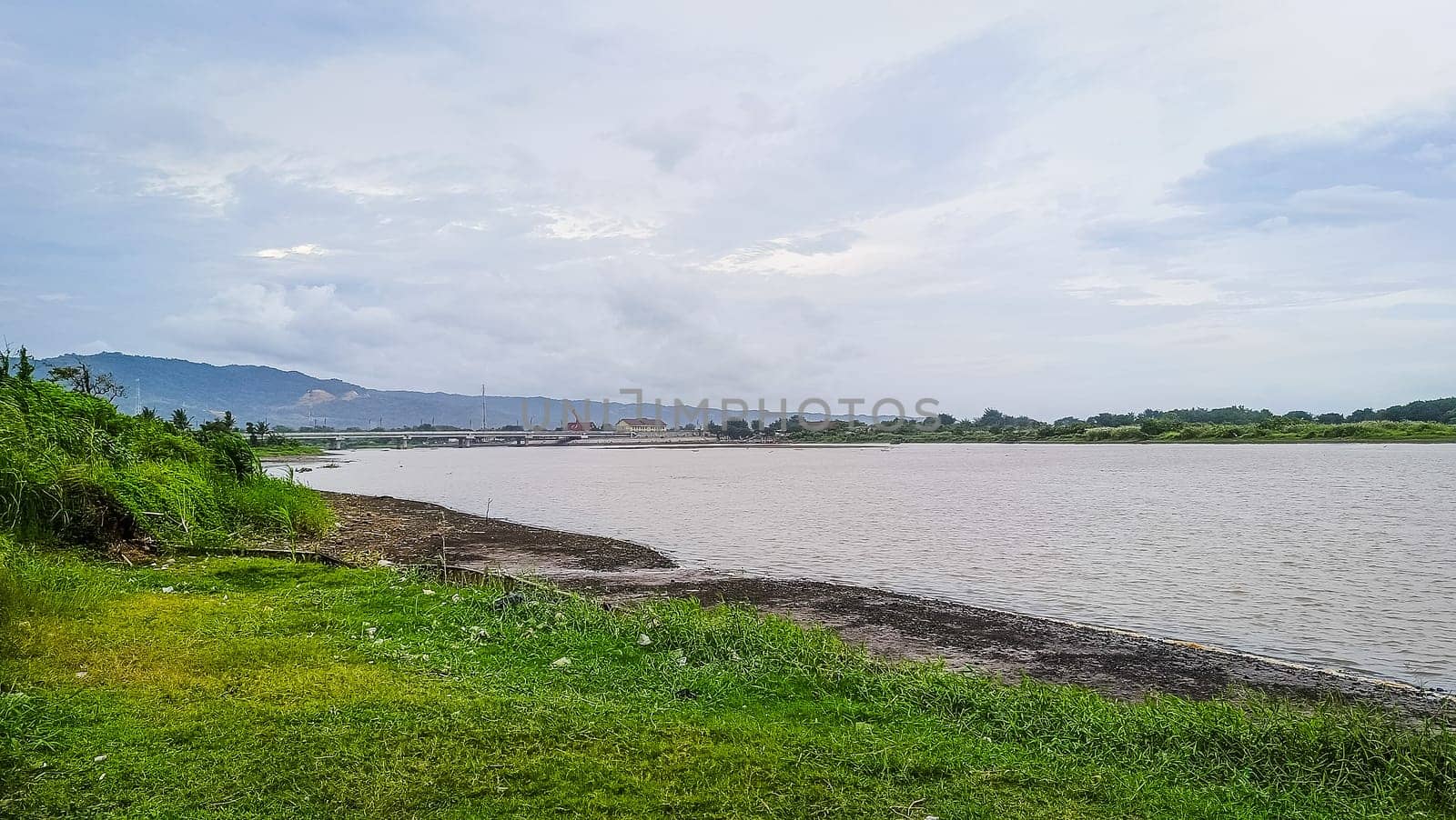 A river with a bridge in the background. The water is calm and the grass is green. The scene is peaceful and serene
