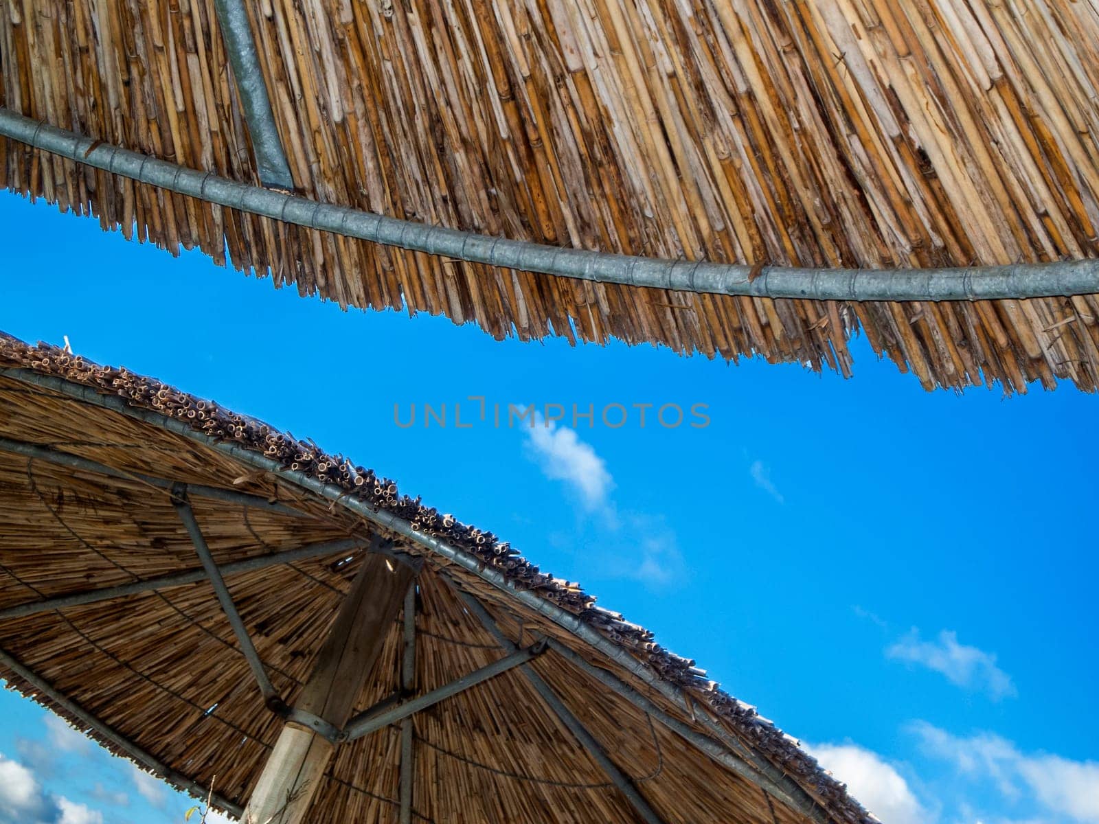 Partial view of the underside of reed covered parasols against a blue sky with scattered clouds.