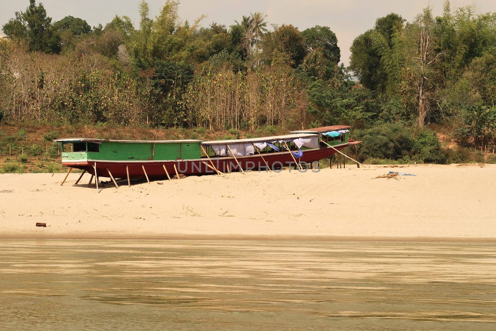 View from the slow boat from Thailand to Laos, another traditional slow boat is jacked up on the beach, shore of the Mekong River, Laos 2023