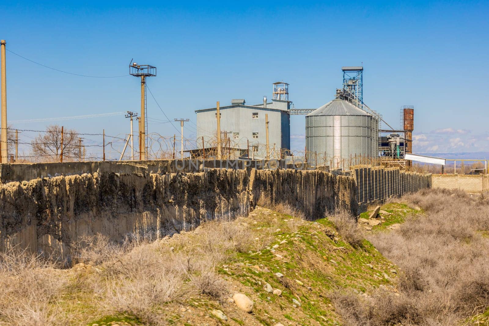 shabby concrete fence with barbwire over and industrial buildings behind at sunny spring day.