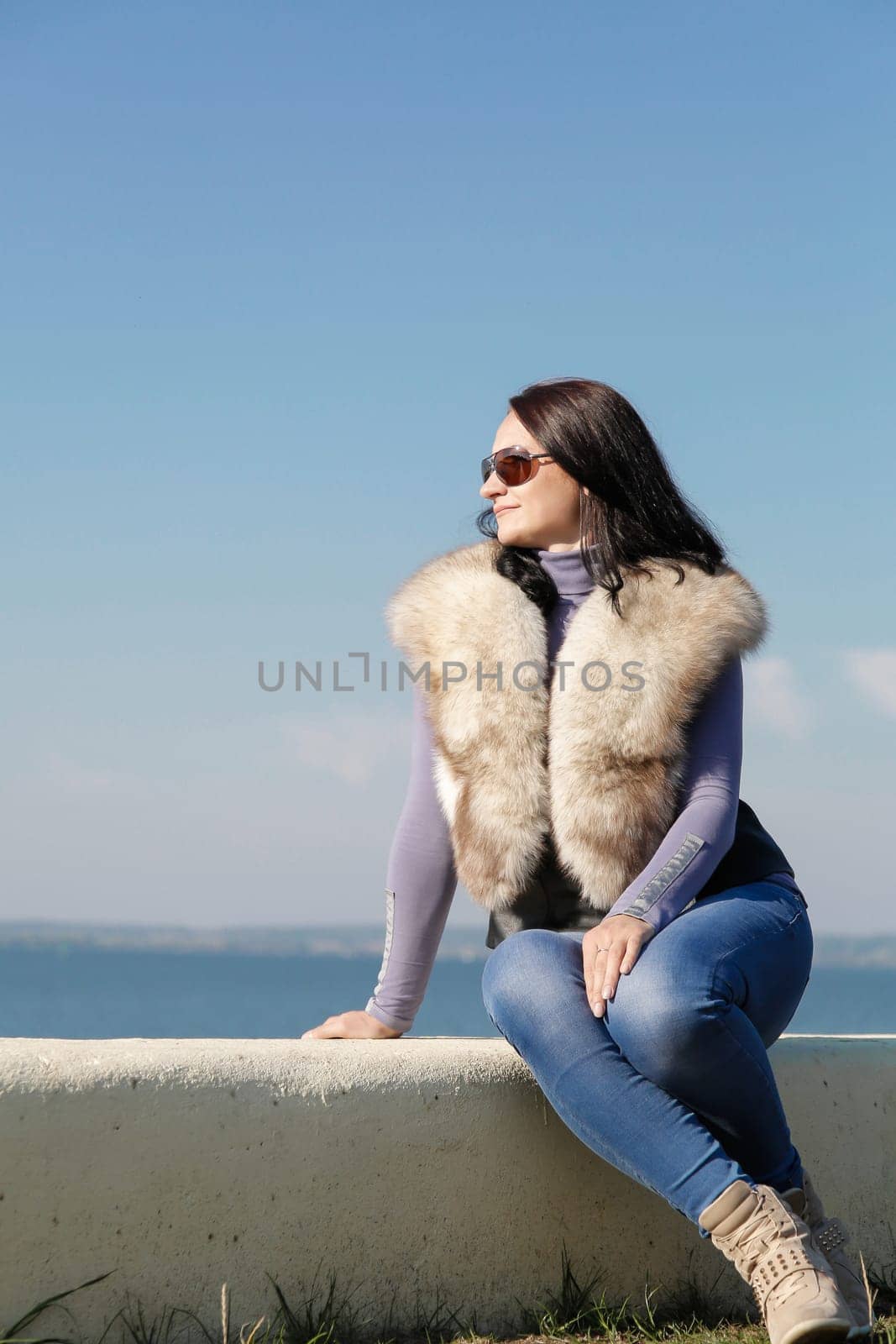 A stylish and fashionable woman in a fur vest sits on a pier against the background of the sea