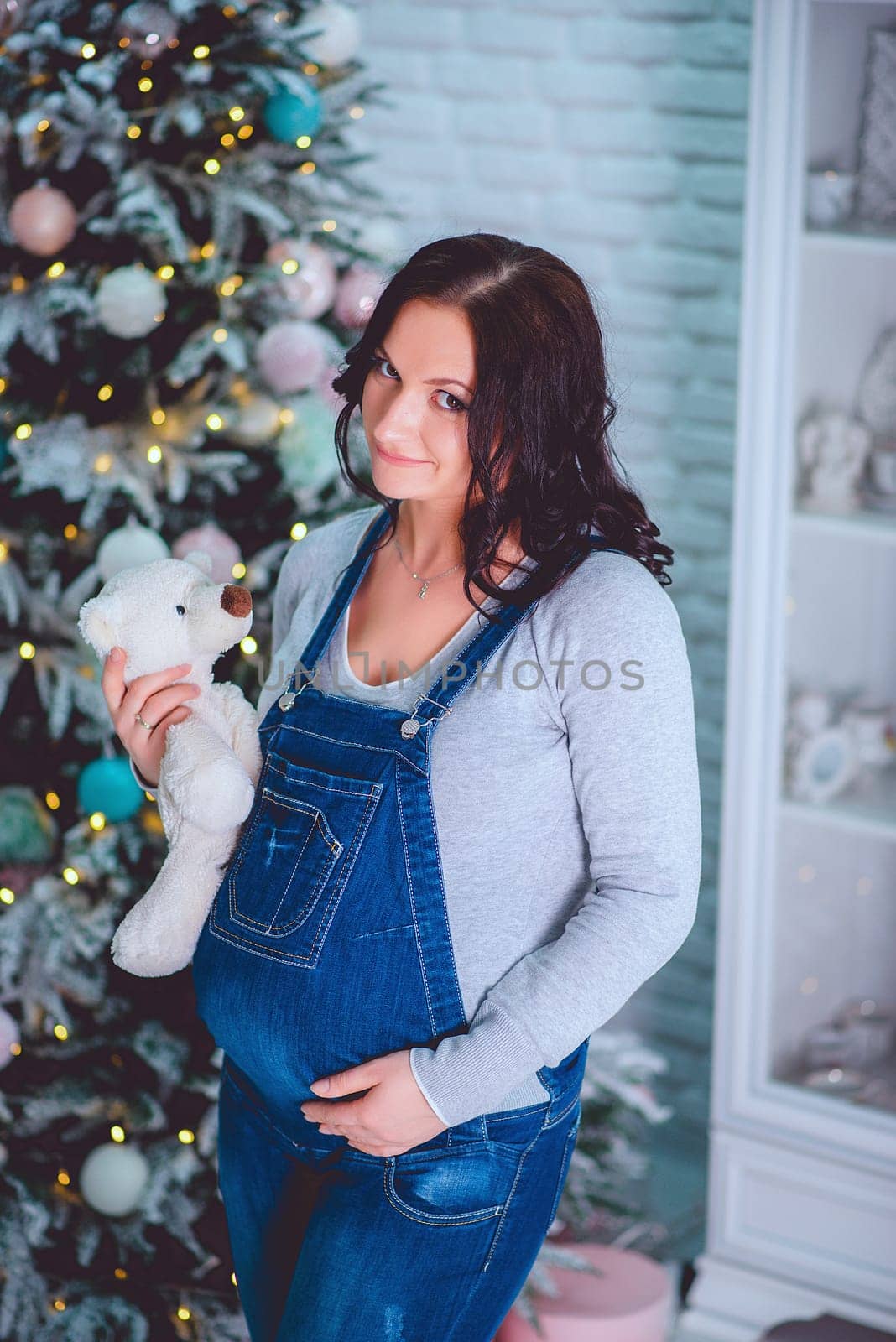 A joyful expectant mother lovingly holding a teddy bear beside a beautifully decorated festive Christmas tree