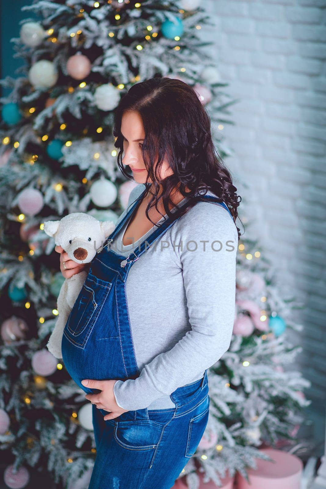 A pregnant girl in a denim jumpsuit with a teddy bear stands near the Christmas tree in the studio