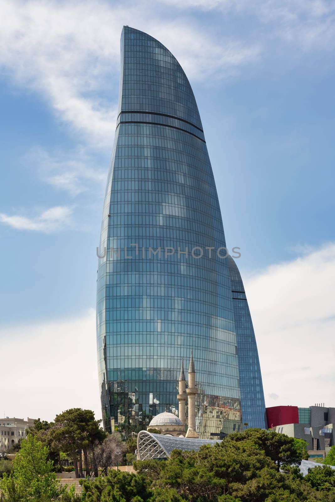 The iconic Flame Towers in Baku, Azerbaijan, rise majestically against a blue sky as seen from Martyrs Lane, resembling flames. Trees in the foreground enhance the cityscape with a natural element