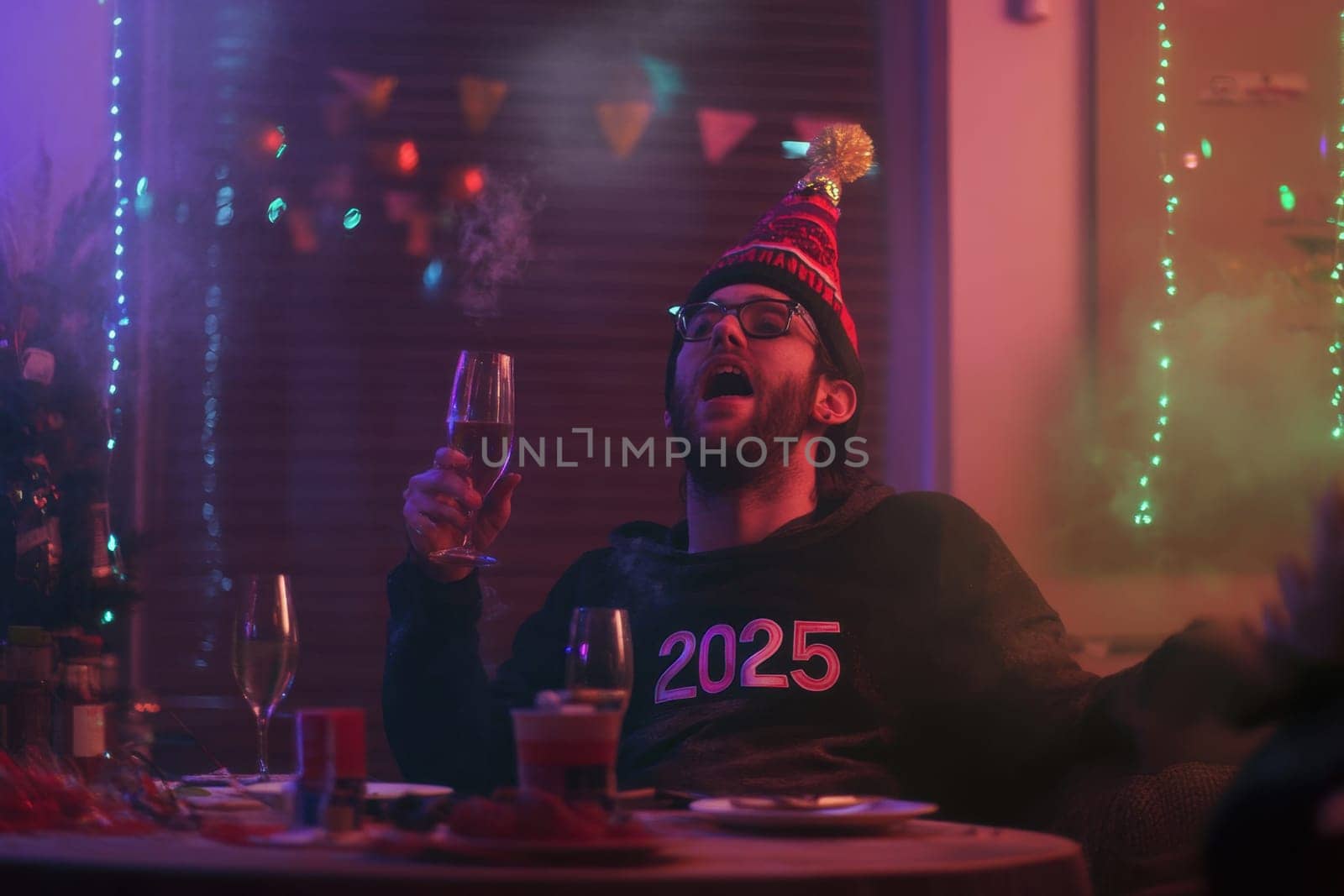 A person wearing a party hat raises a glass in celebration as colorful lights illuminate the space ushering in 2025.