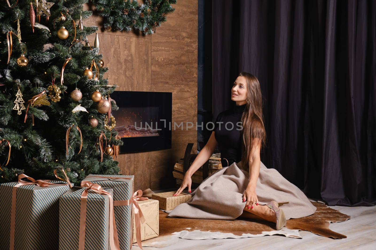 A dreamy woman sits on the floor beside a warm fireplace and looking at Christmas tree decorated with toys and balls