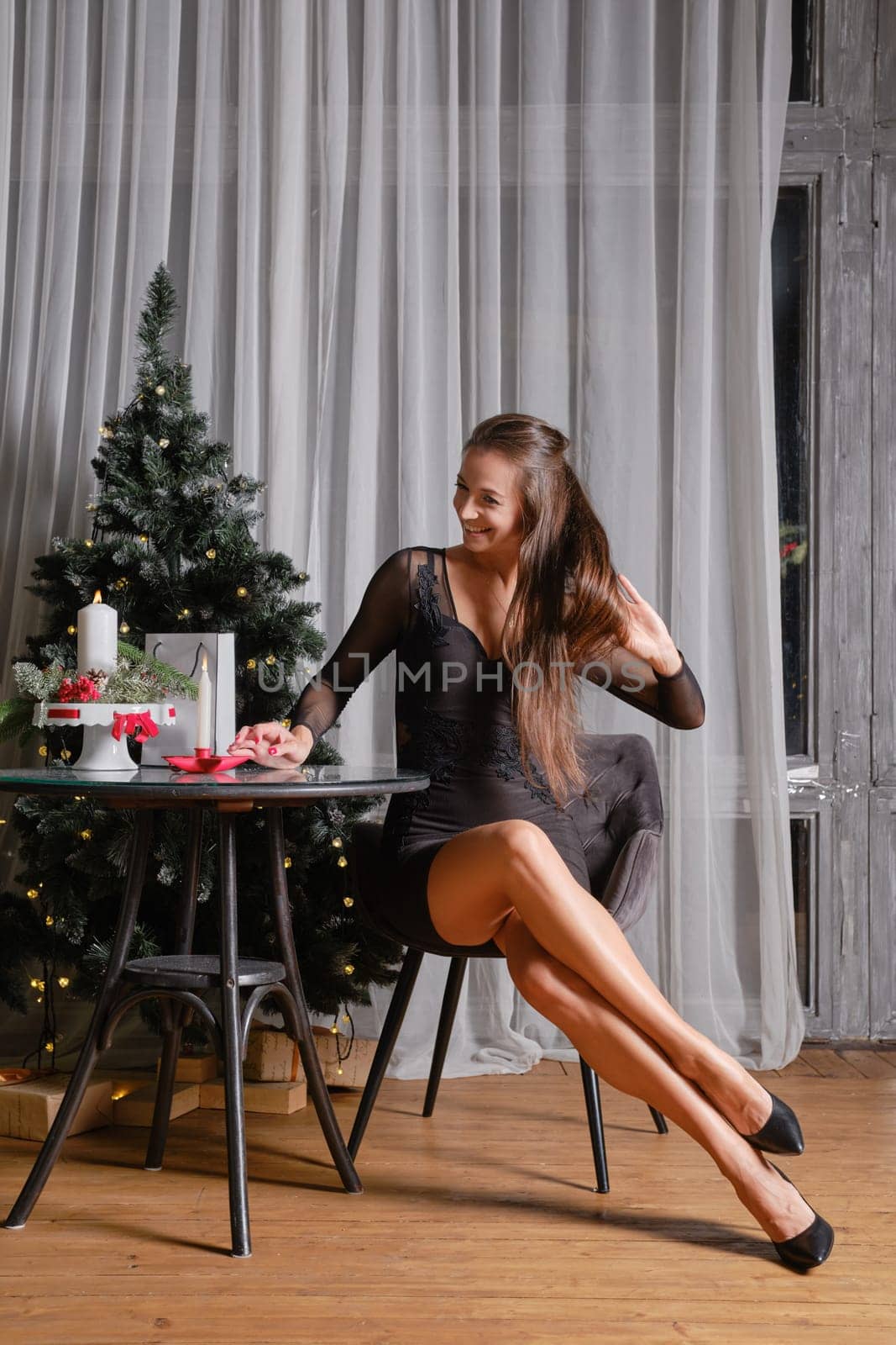 A stylish woman in a tight black dress sits beside a Christmas tree and touching her long hair