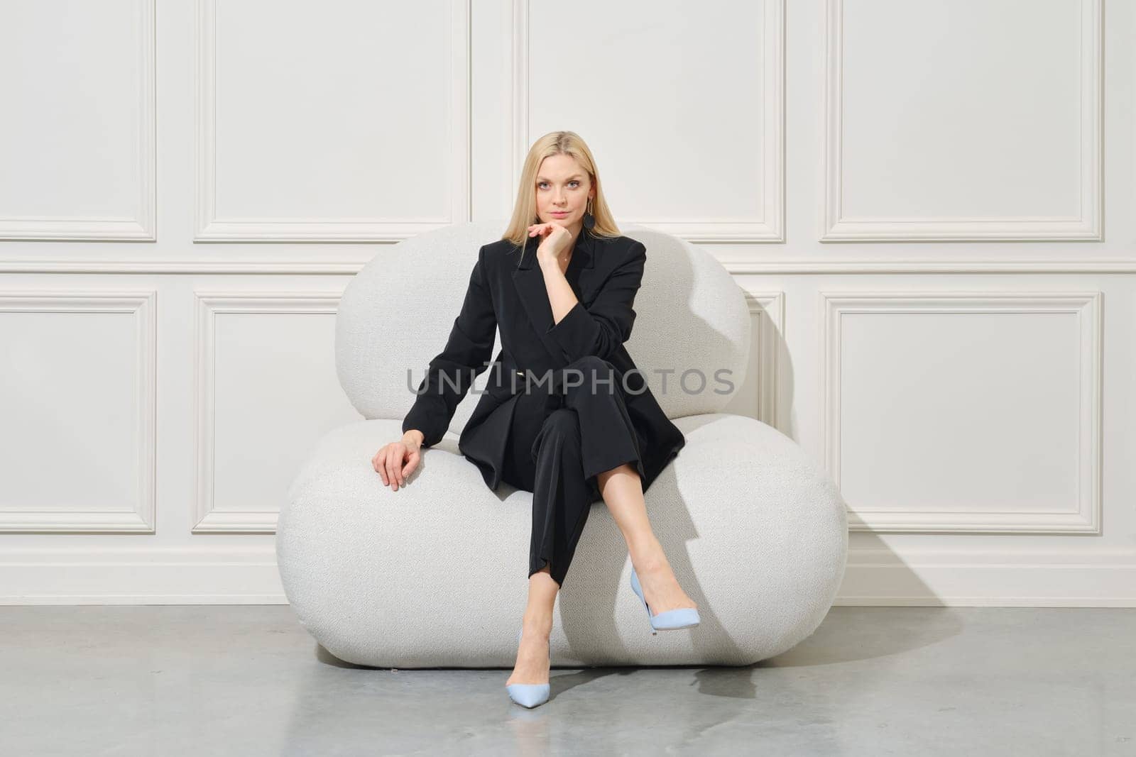 Positive businesswoman in casual suit sits on large bean bag propping chin and looking with interest