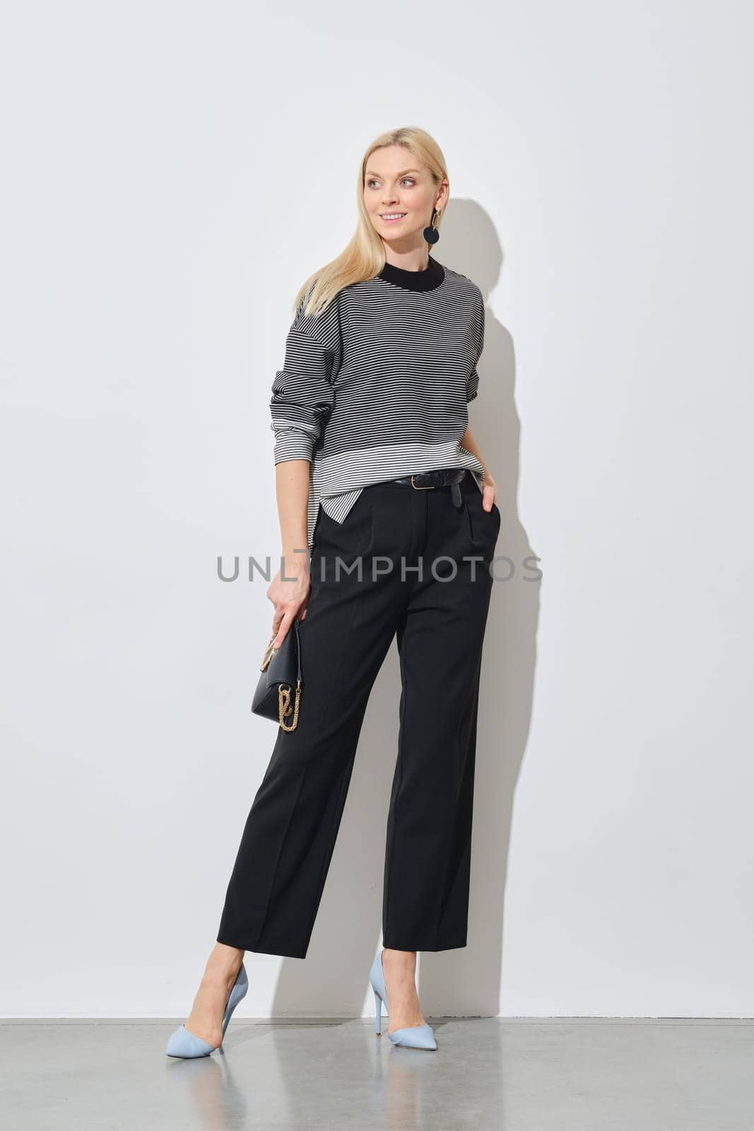 A fashionable woman poses in a striped top and black trousers, accessorized with blue heels and a small purse against a white wall