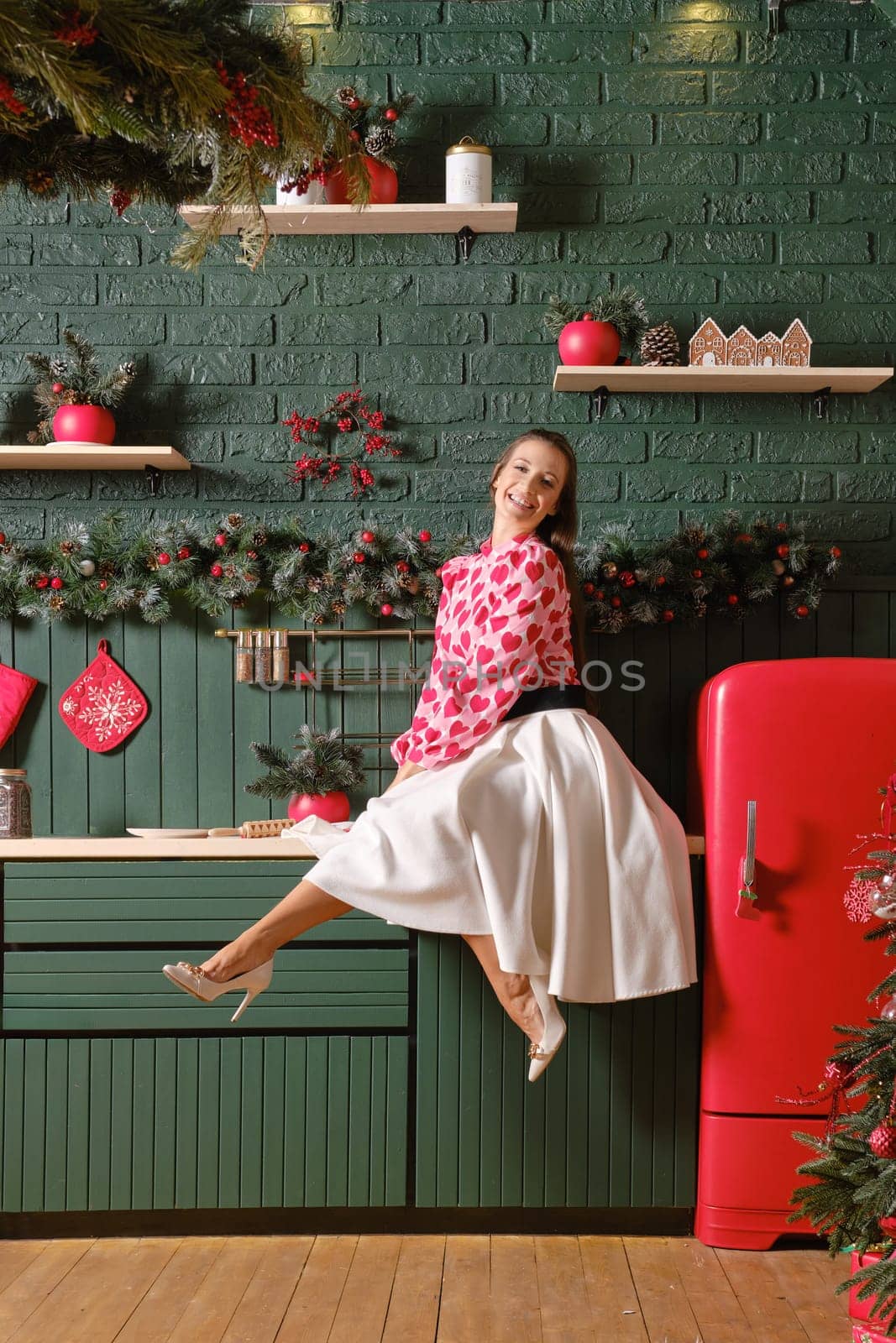 Laughing woman sits in a festive mood in a kitchen decorated for the Christmas season. She wears a white skirt and a red top with heart patterns, complementing the green and red decor around her.