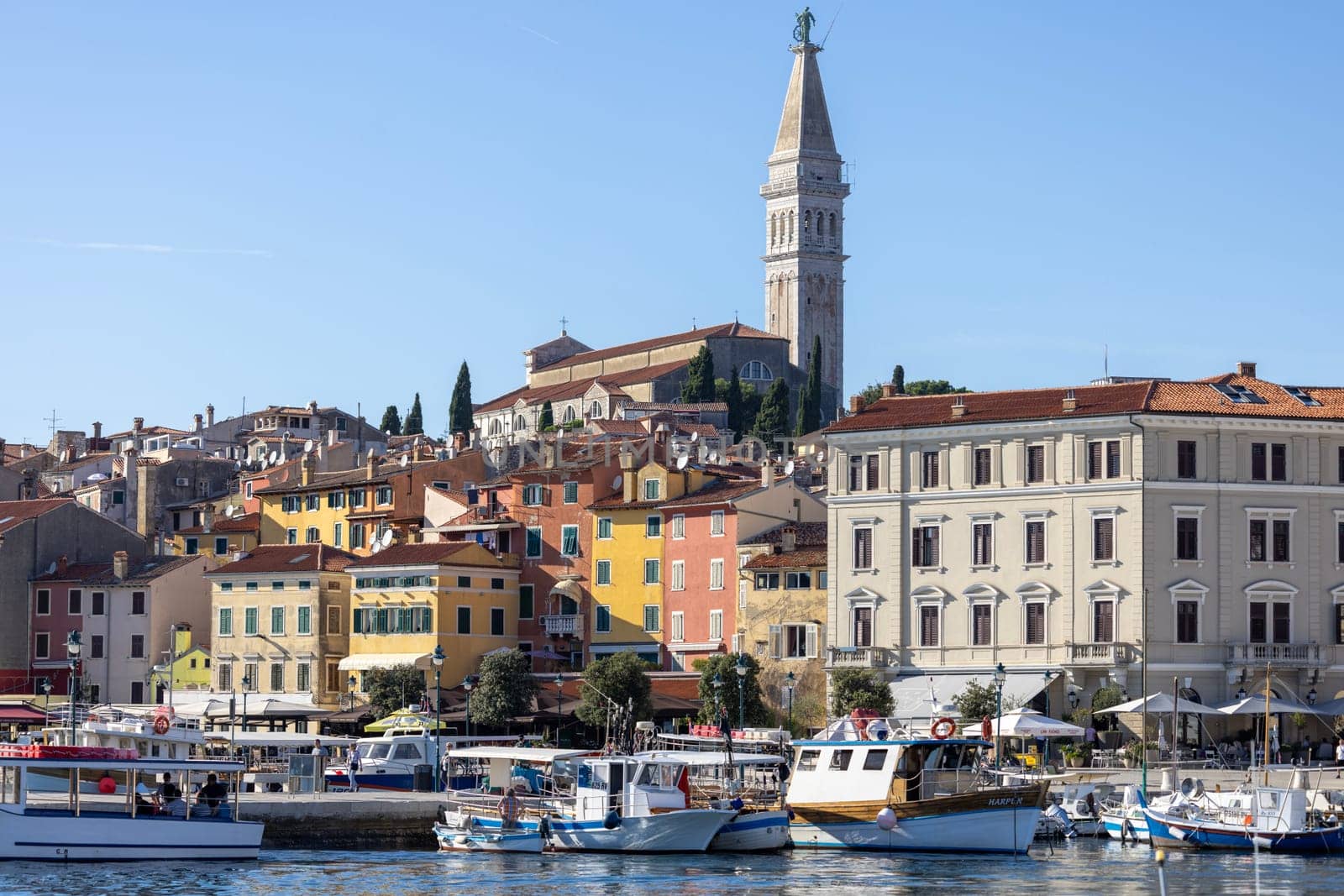 Rovinj, Croatia, Istria - September 29, 2023: Colorful tenement houses on the boulevard by South port over Adriatic Sea, moored boats and Saint Euphemia Church in a distance