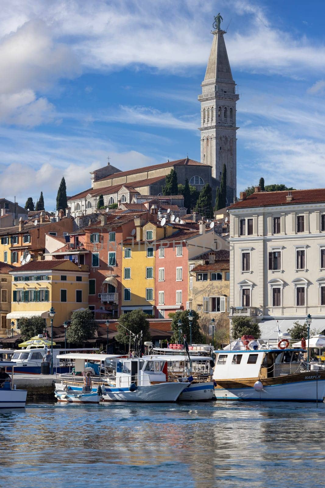 Rovinj, Croatia, Istria - September 29, 2023: Colorful tenement houses on the boulevard by South port over Adriatic Sea, moored boats and Saint Euphemia Church in a distance