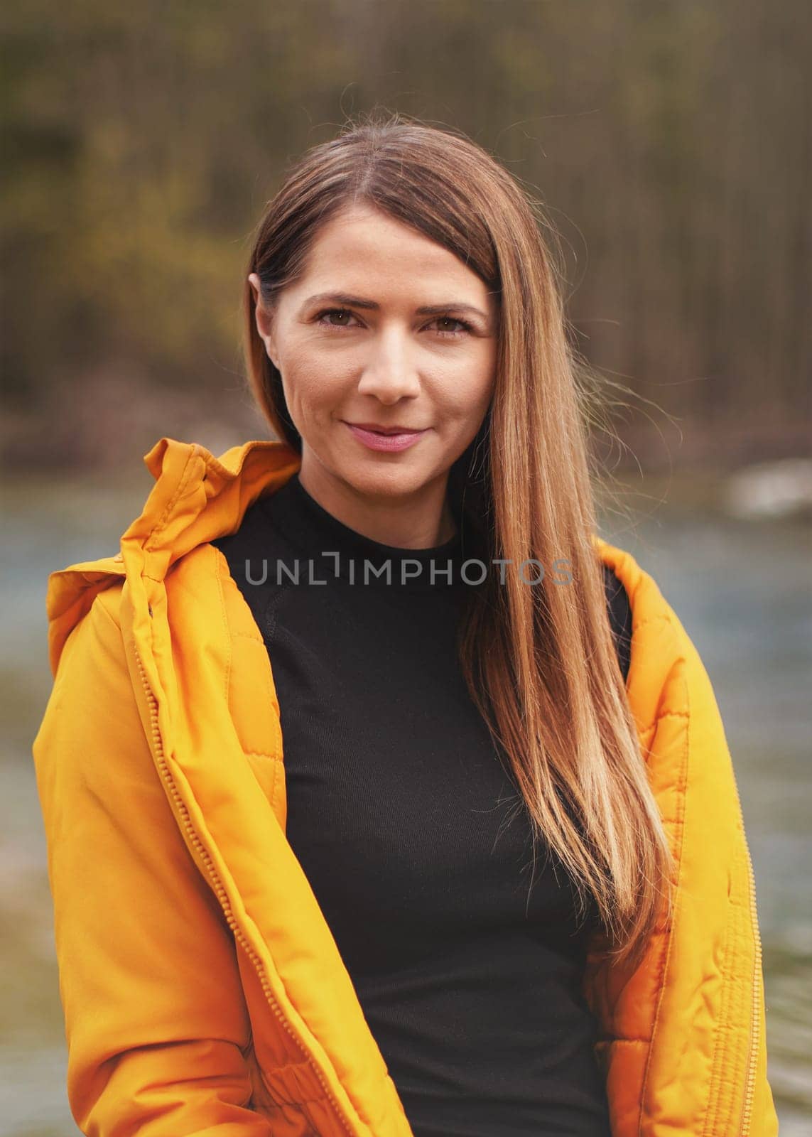 Portrait of young woman in yellow jacket, smiling, long hair down, blurred trees and river background