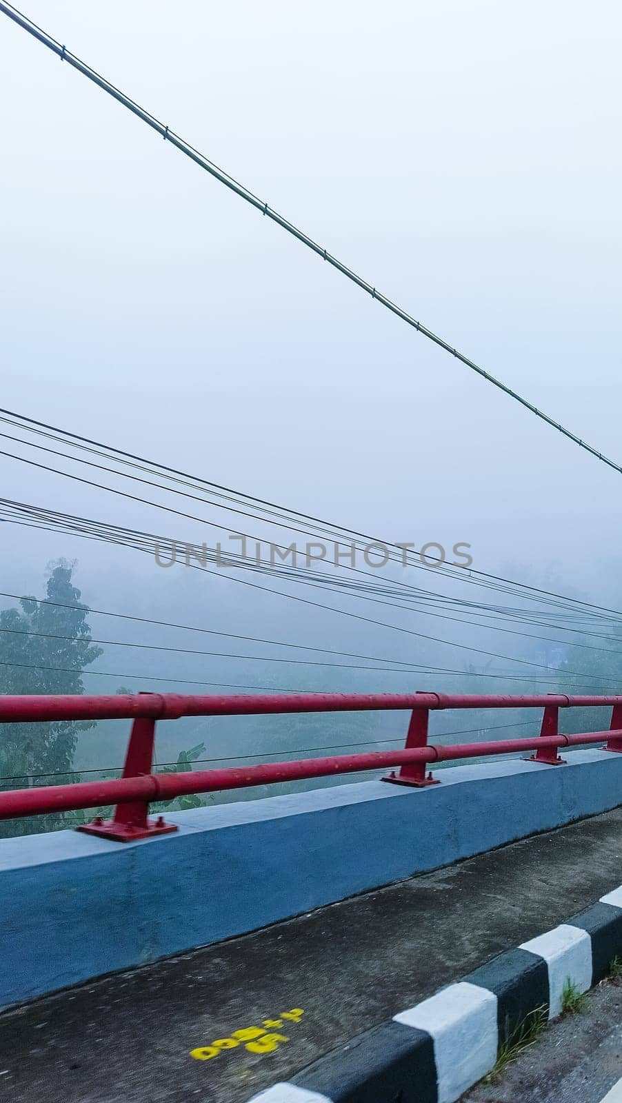 A picturesque scene of a foggy landscape viewed from a bridge. Red railings and power lines create a sense of mystery and tranquility in this ethereal setting.