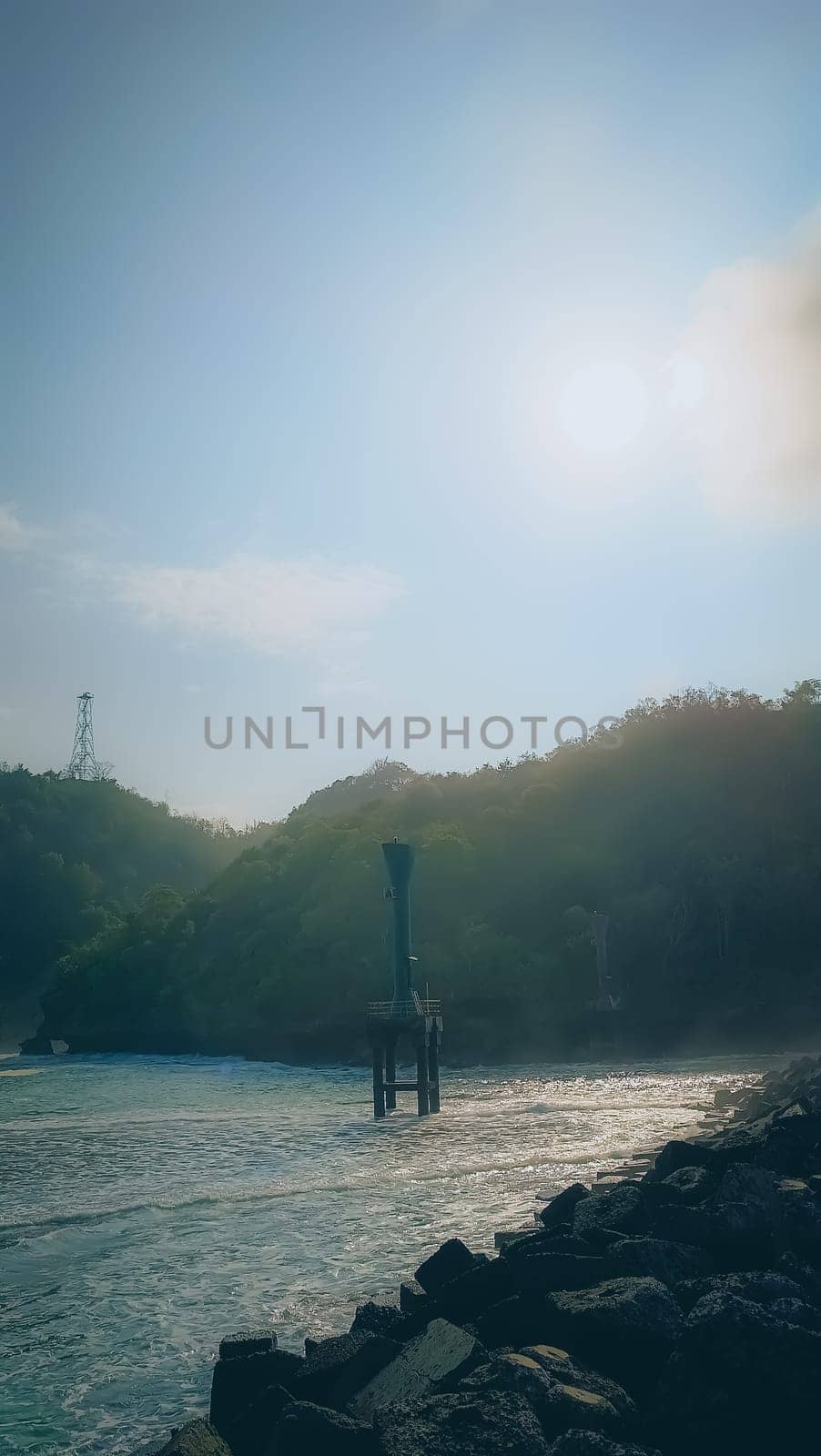 A rocky shoreline with a large rock formation in the background. The water is calm and the sky is blue