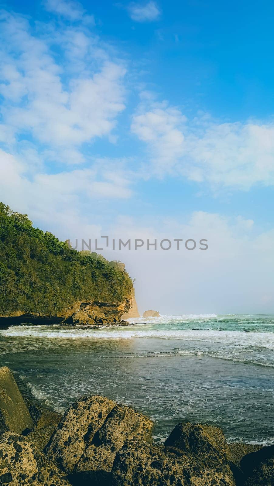 A rocky shoreline with a large rock formation in the background. The water is calm and the sky is blue