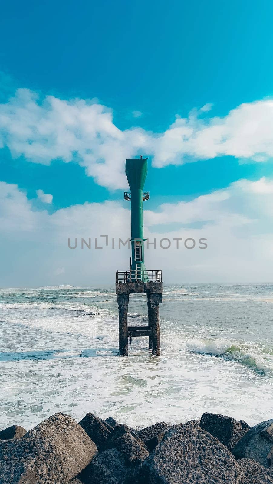 A solitary beacon tower stands on rocky coast, surrounded by the ocean under a clear blue sky. Perfect for themes of solitude, navigation, and coastal landscapes.