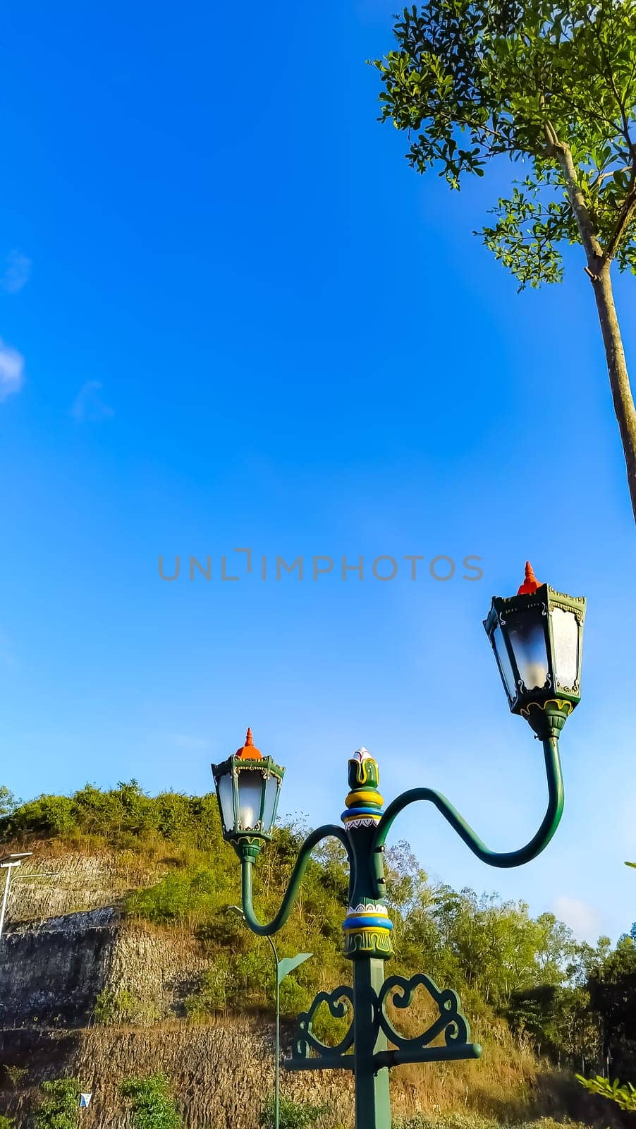 Elegant green street lamps with intricate designs stand in front of a vivid blue sky and lush hill. The scene exudes a blend of nature and historical architecture.