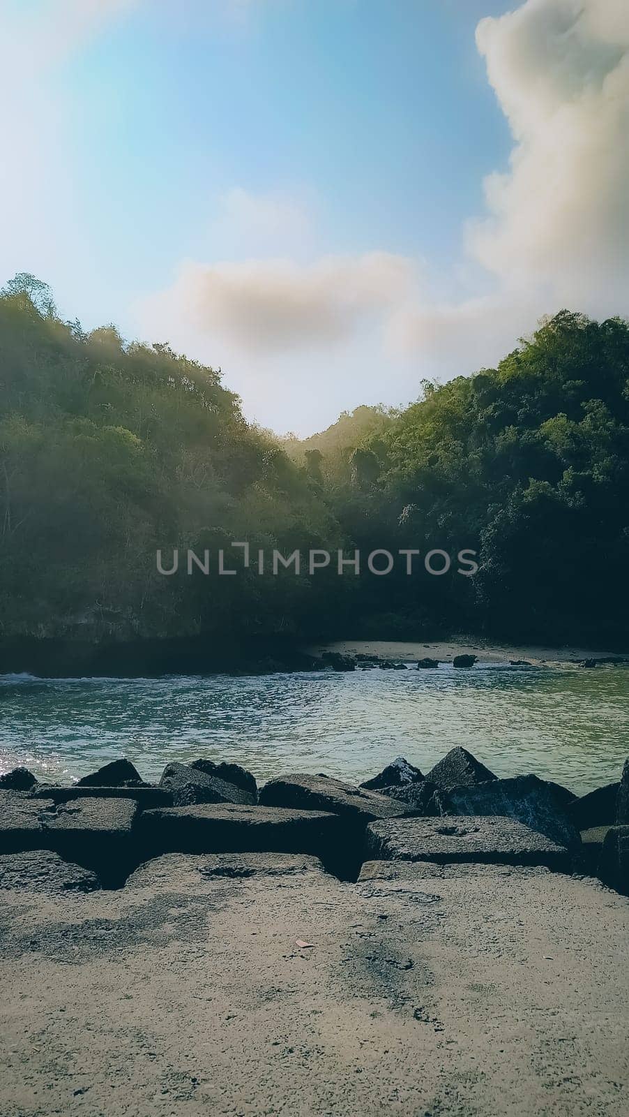 A rocky shoreline with a large rock formation in the background. The water is calm and the sky is blue