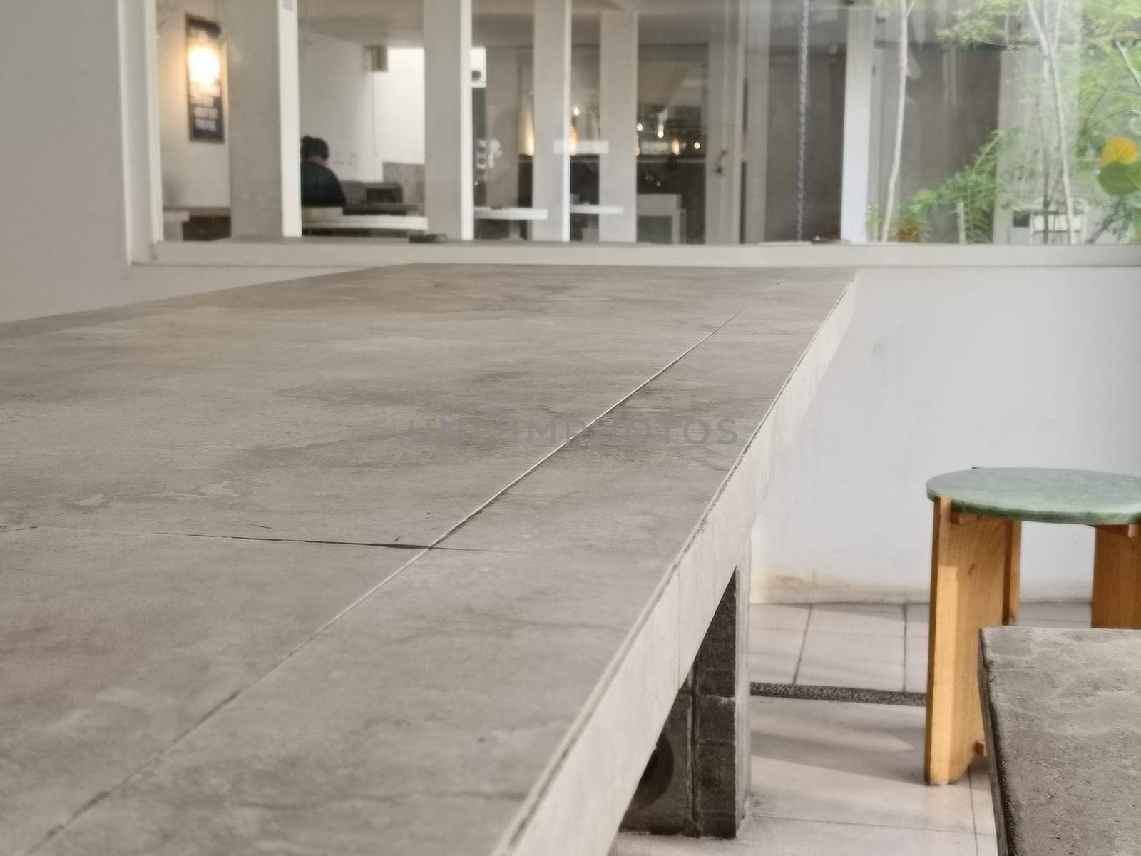 A modern minimalist cafe interior featuring a large concrete table and greenery. The bright space is filled with natural light, providing a serene and inviting atmosphere.