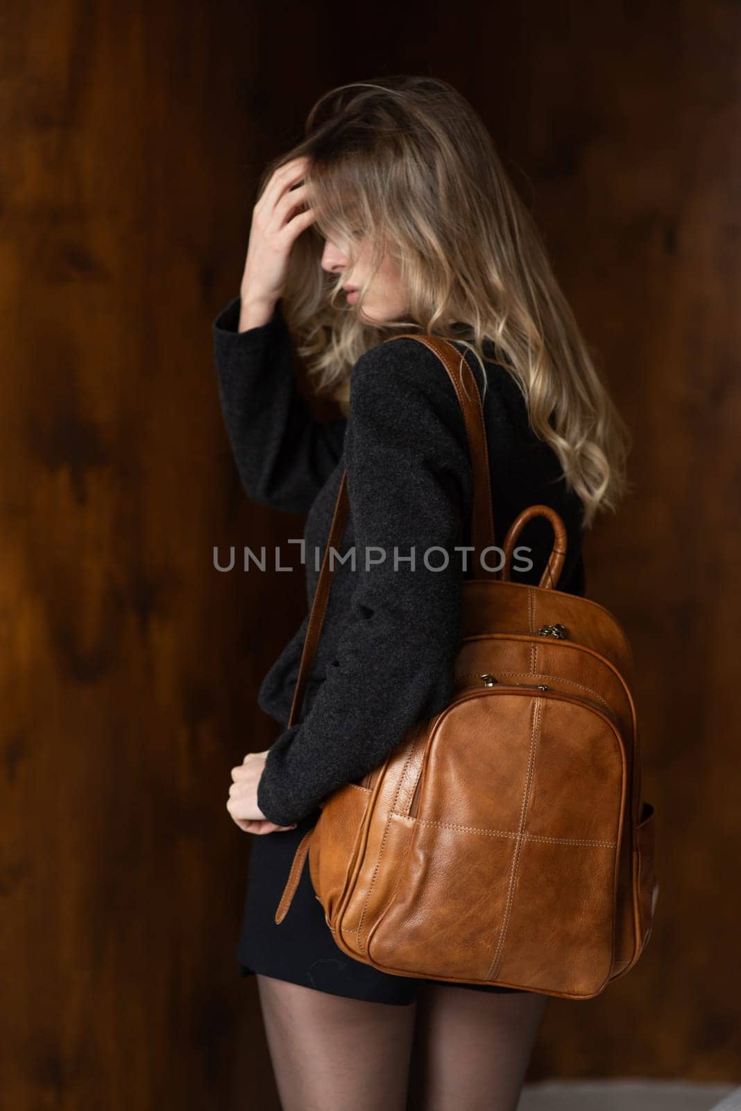 A girl in a warm sweater, a black short skirt and nylon tights is posing with a light brown backpack