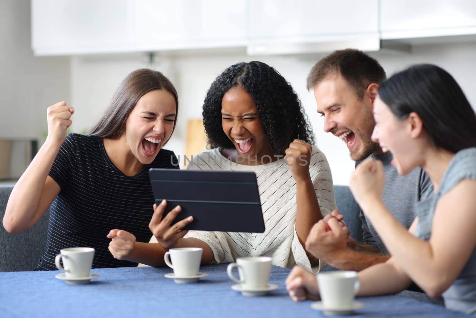 Four excited interracial friends watching media on tablet in the kitchen at home