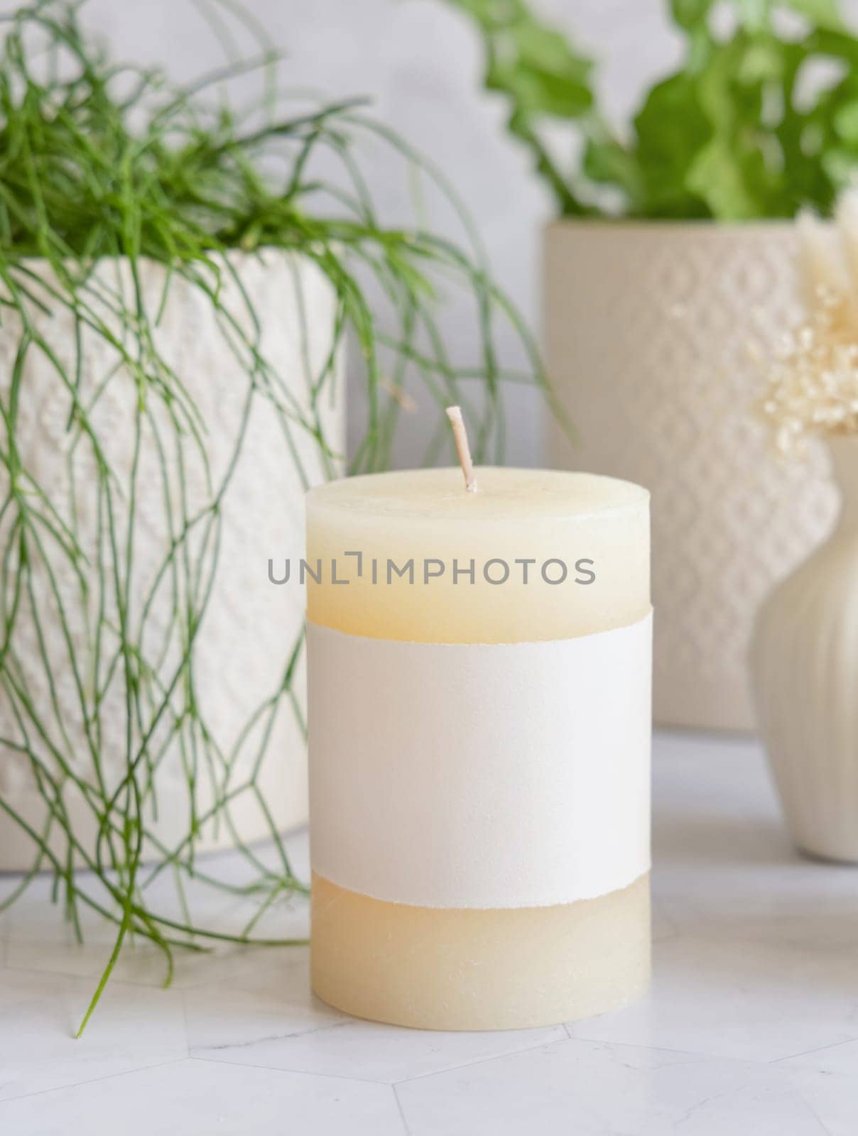 Pillar candle with blank label on marble near potted green plants close up, packaging mockup. Minimal scandinavian home decoration, romantic interior