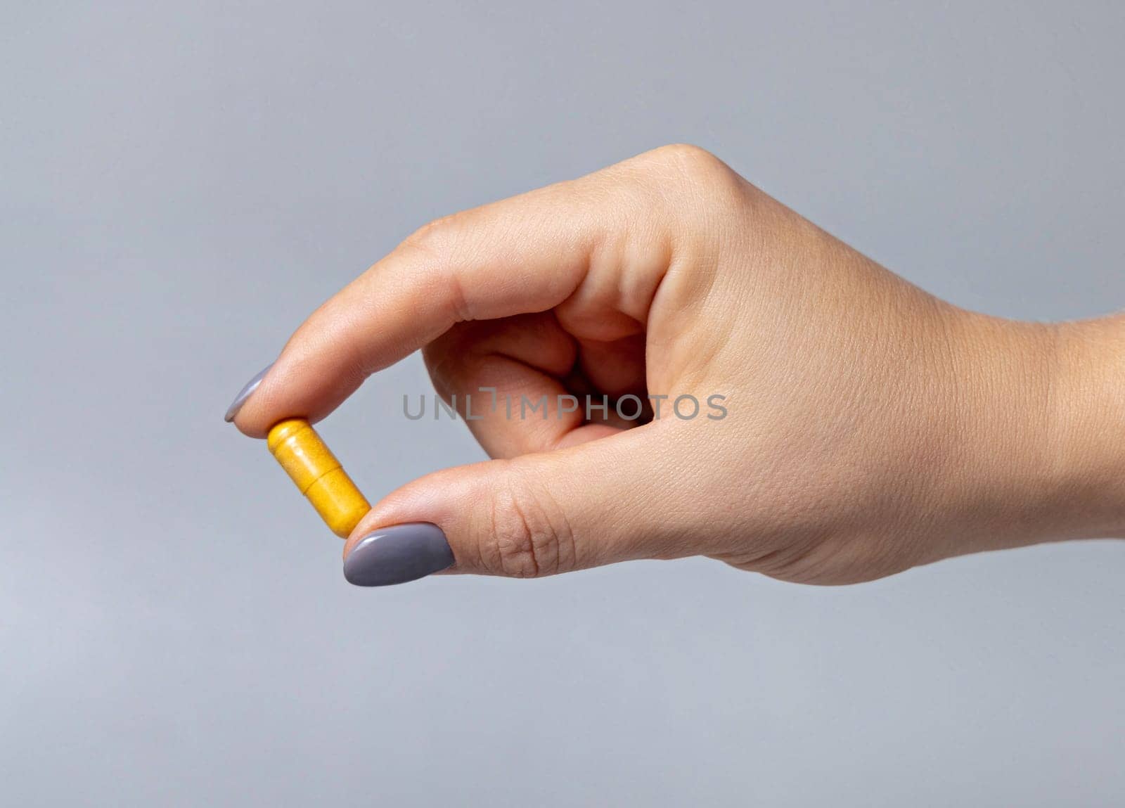 Female hand holding a yellow capsule with fingers closeup against grey background. Healthcare, medical and pharmaceutical concept.