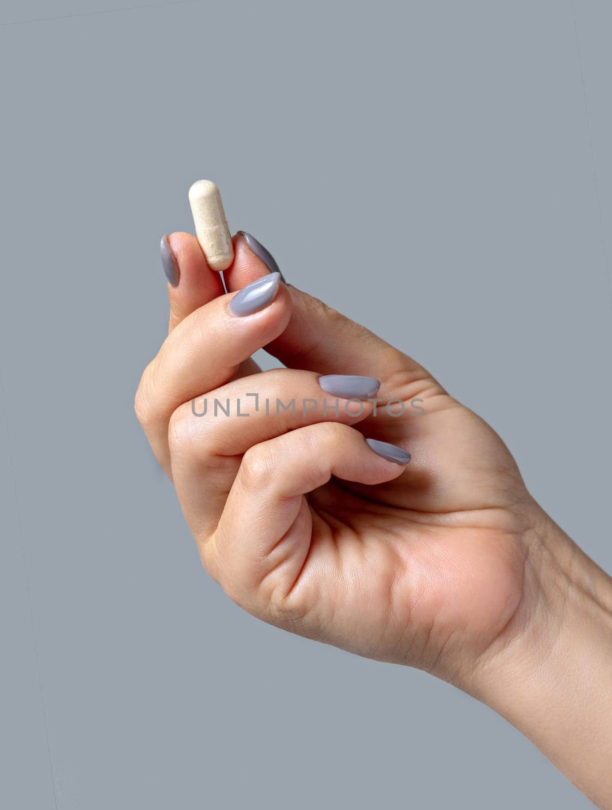 Female hand holding a beige capsule with fingers closeup against grey background. Healthcare, medical and pharmaceutical concept
