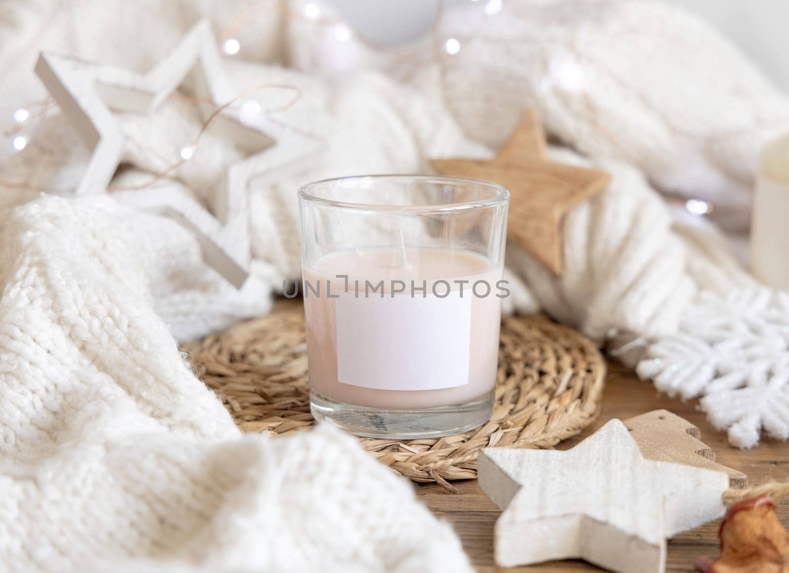 Glass candle with blank label near white knitted sweater, wooden Christmas decor and lights, copy space. Winter atmospheric mock up in rustic or scandinavian style