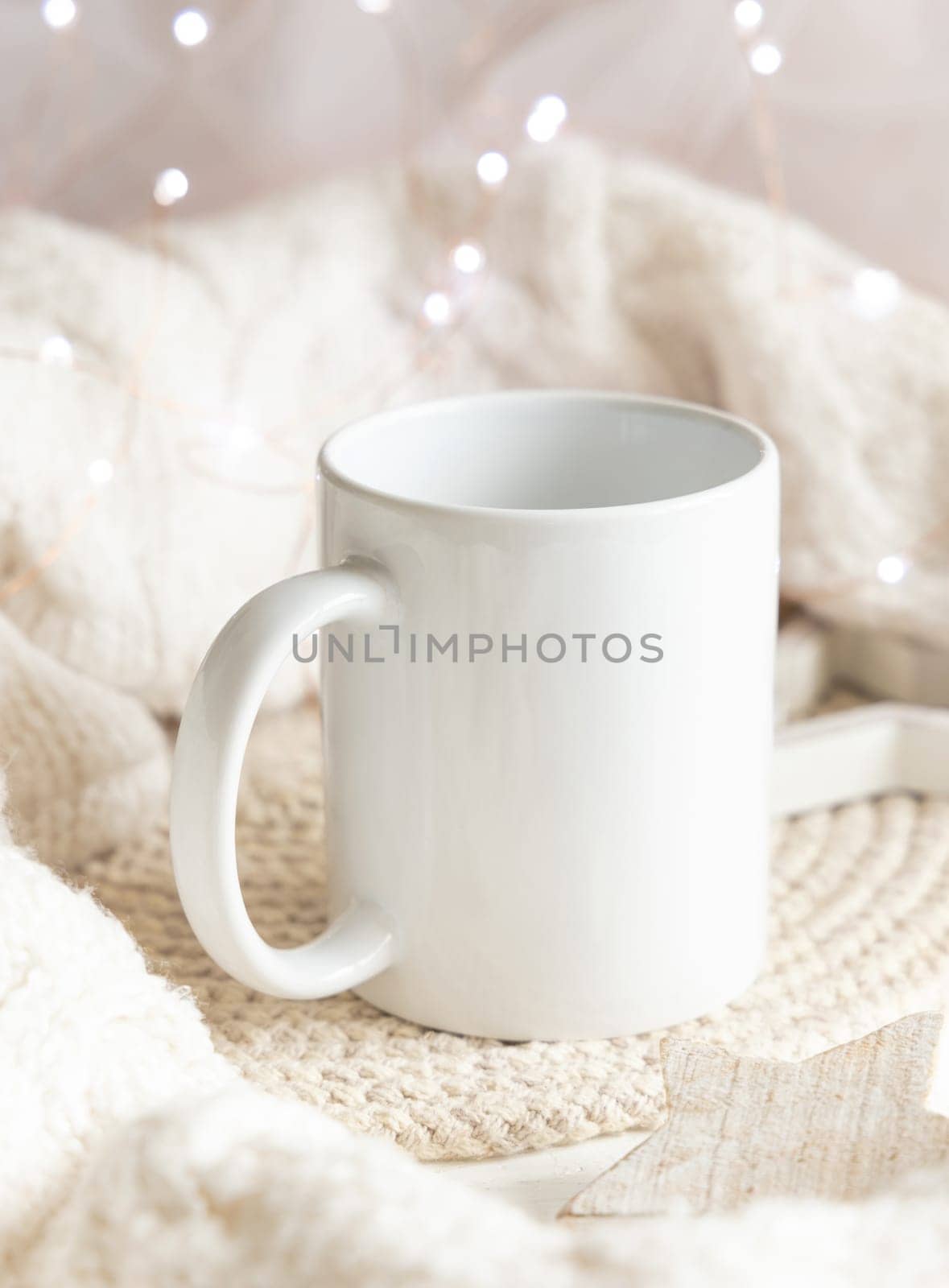 White blank coffee mug near white knitted sweater, wooden Christmas decor and lights, copy space. Winter atmospheric mock up in rustic or scandinavian style