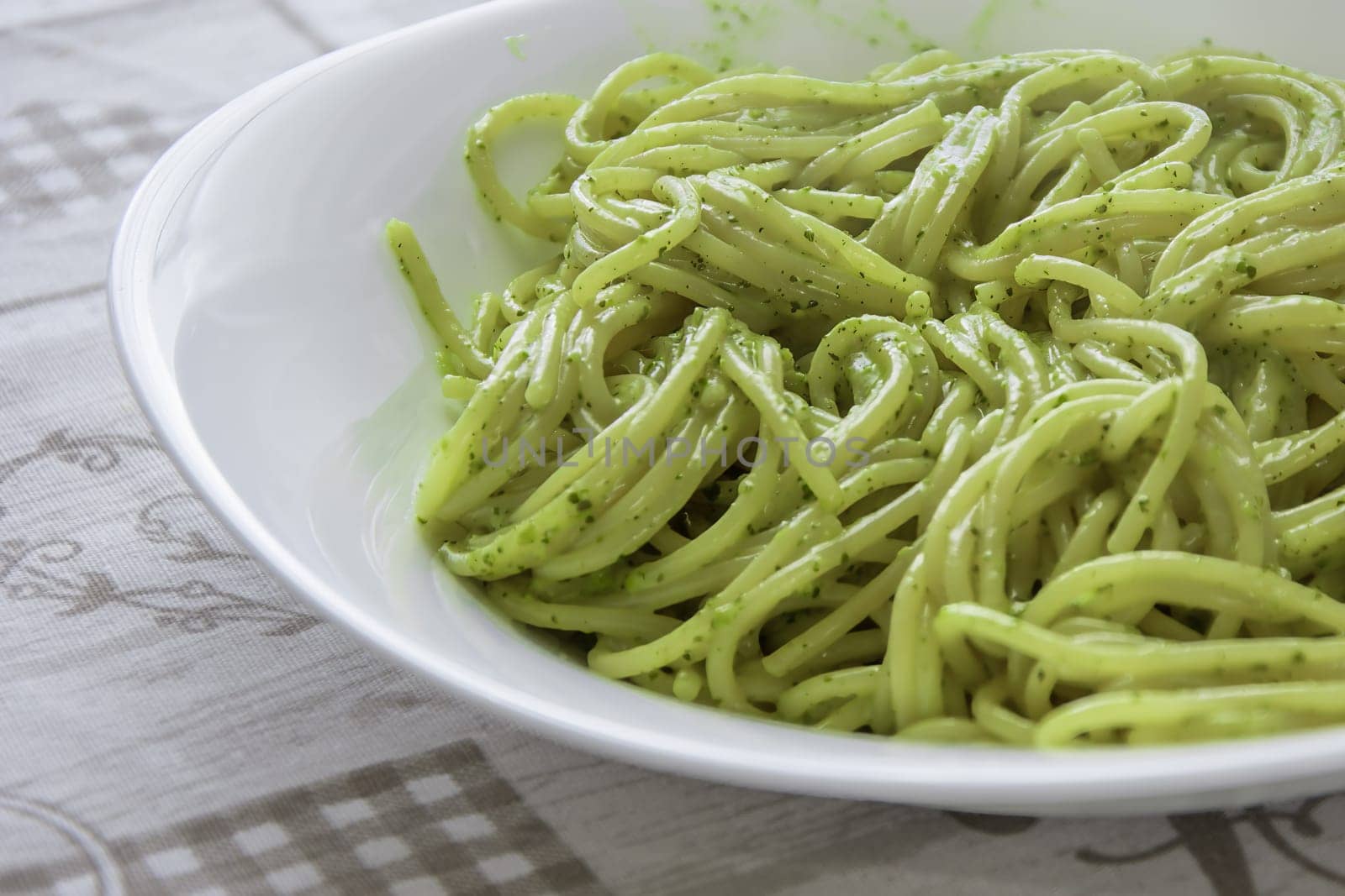 A close-up shot of spaghetti with traditional Genovese pesto sauce, served in a white bowl on a patterned tablecloth. Ideal for concepts of Italian cuisine, traditional recipes, and Mediterranean flavors.pasta, spaghetti, pesto, Italian food, dish, meal, cuisine, bowl, food, plate