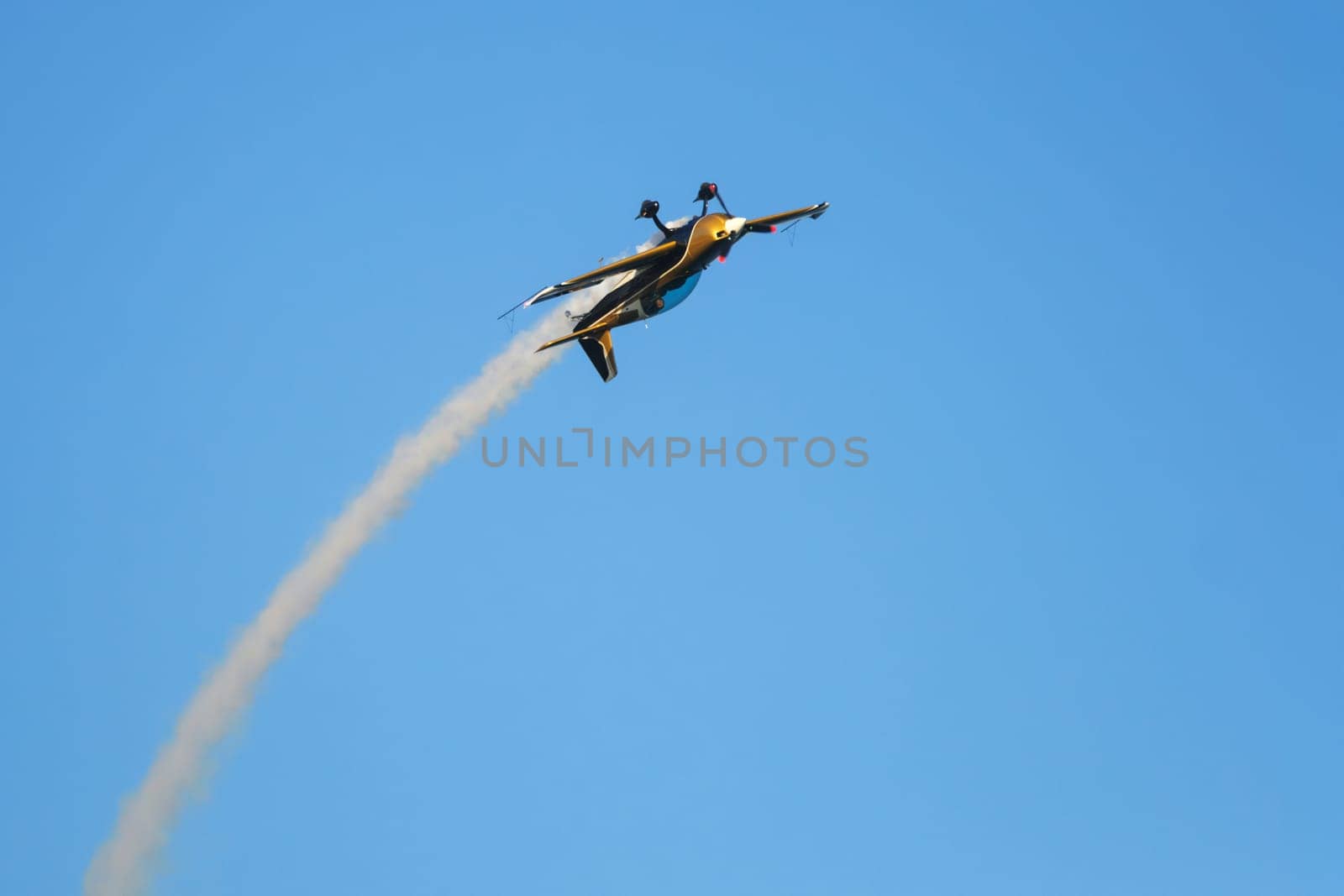 Golden stunt aerobatic plane inverted flying performing stunts in blue sky with smoke trail. Concept of speed, difficulty, precision, risk