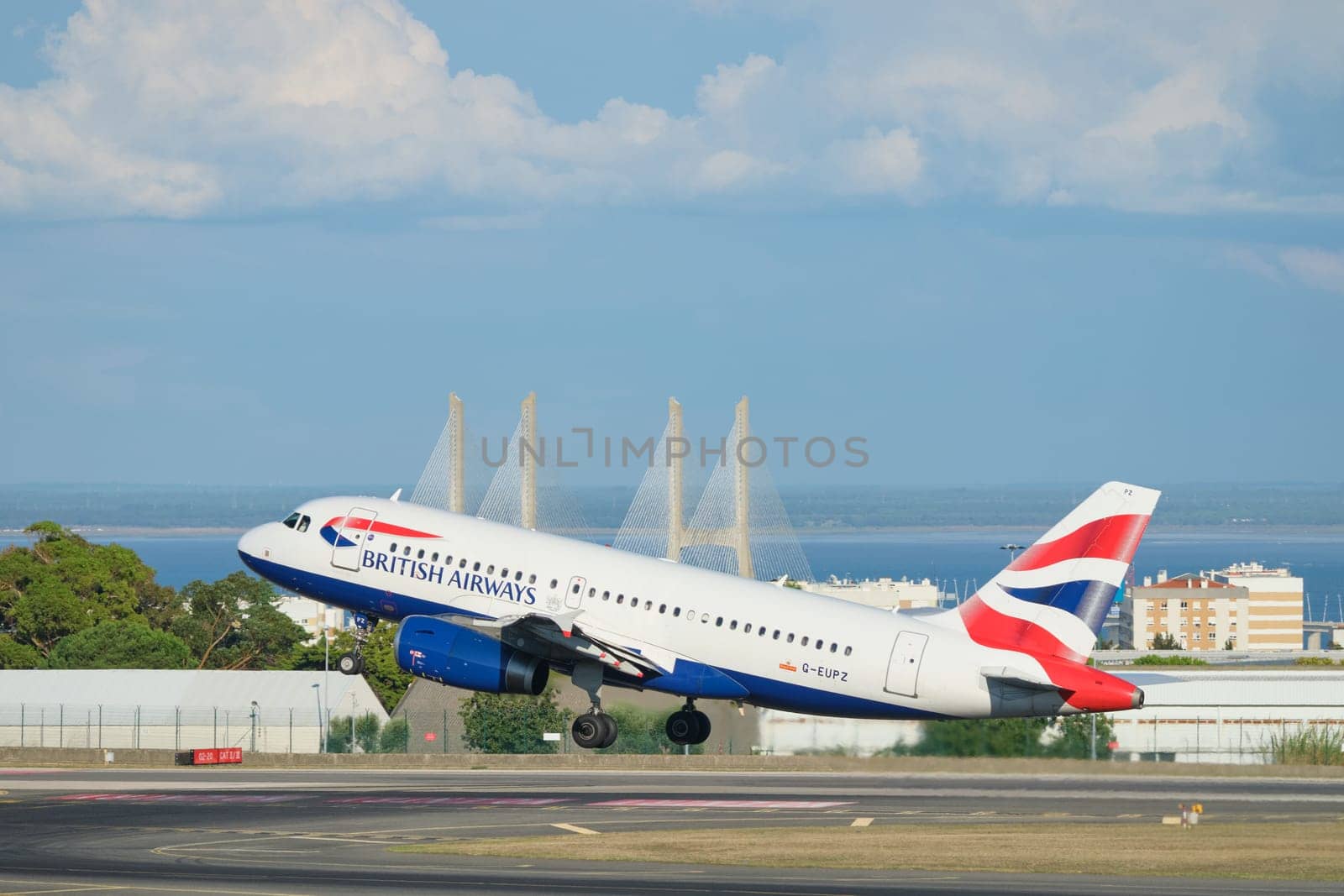 Lisbon, Portugal - September 2, 2023: British Airways Airbus A319-131 passenger plane take off in Humberto Delgado Airport in Lisbon with Vasco da Gamma bridge in background