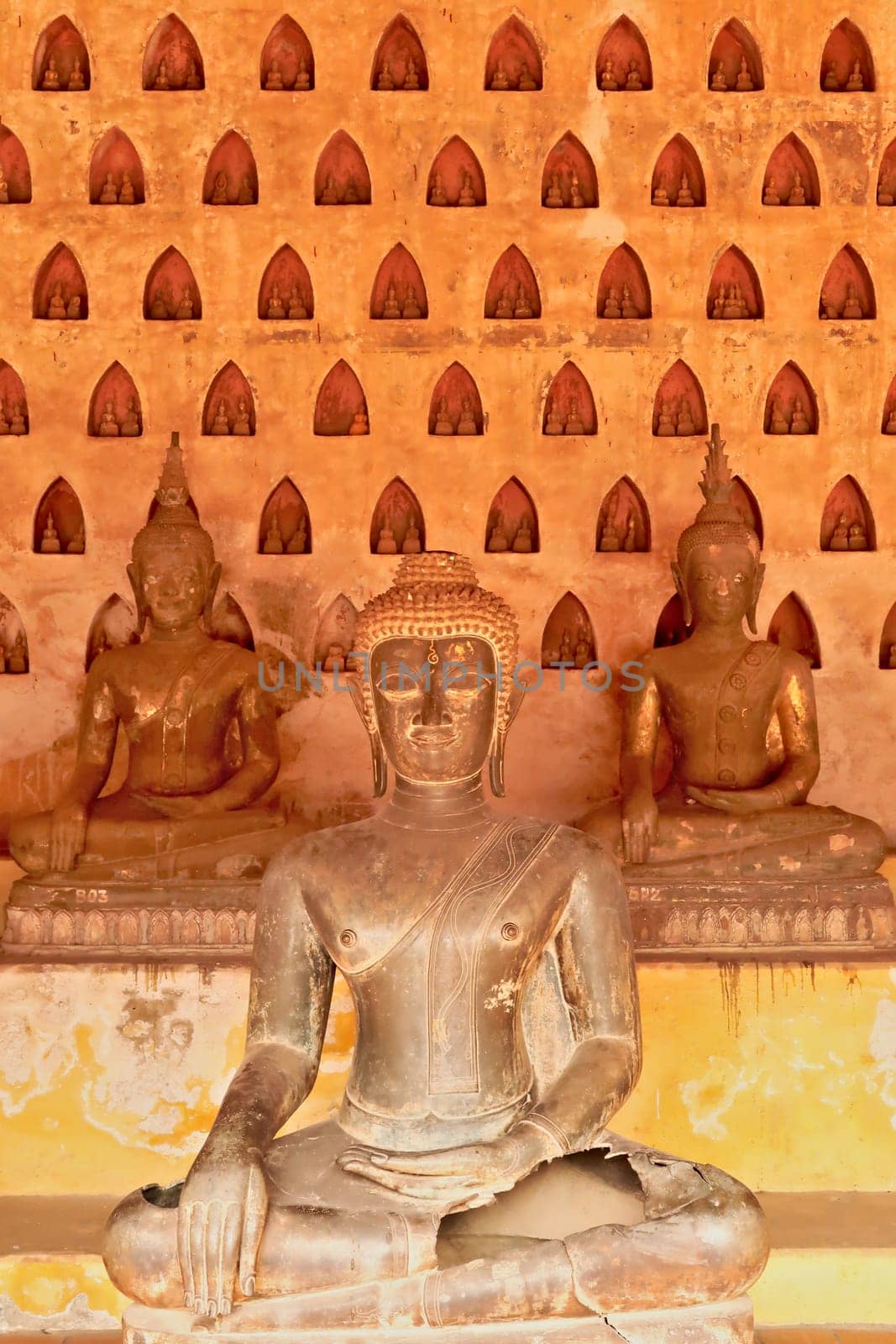 Three big buddha statues in front of a wall filled with many miniature buddha figurines in the cloister wall of Wat Si Saket, a buddhist temple in Vientiane, Laos 2023