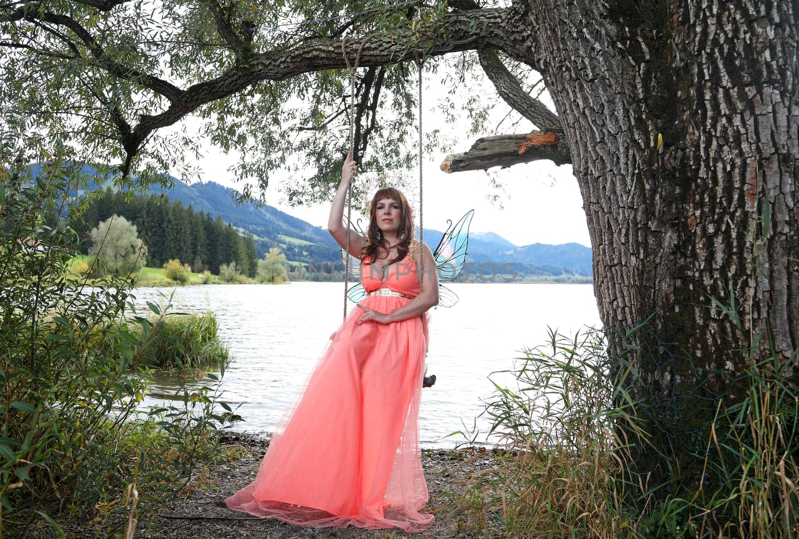 A beautiful elf with a crown and wings sits on a swing by the lake