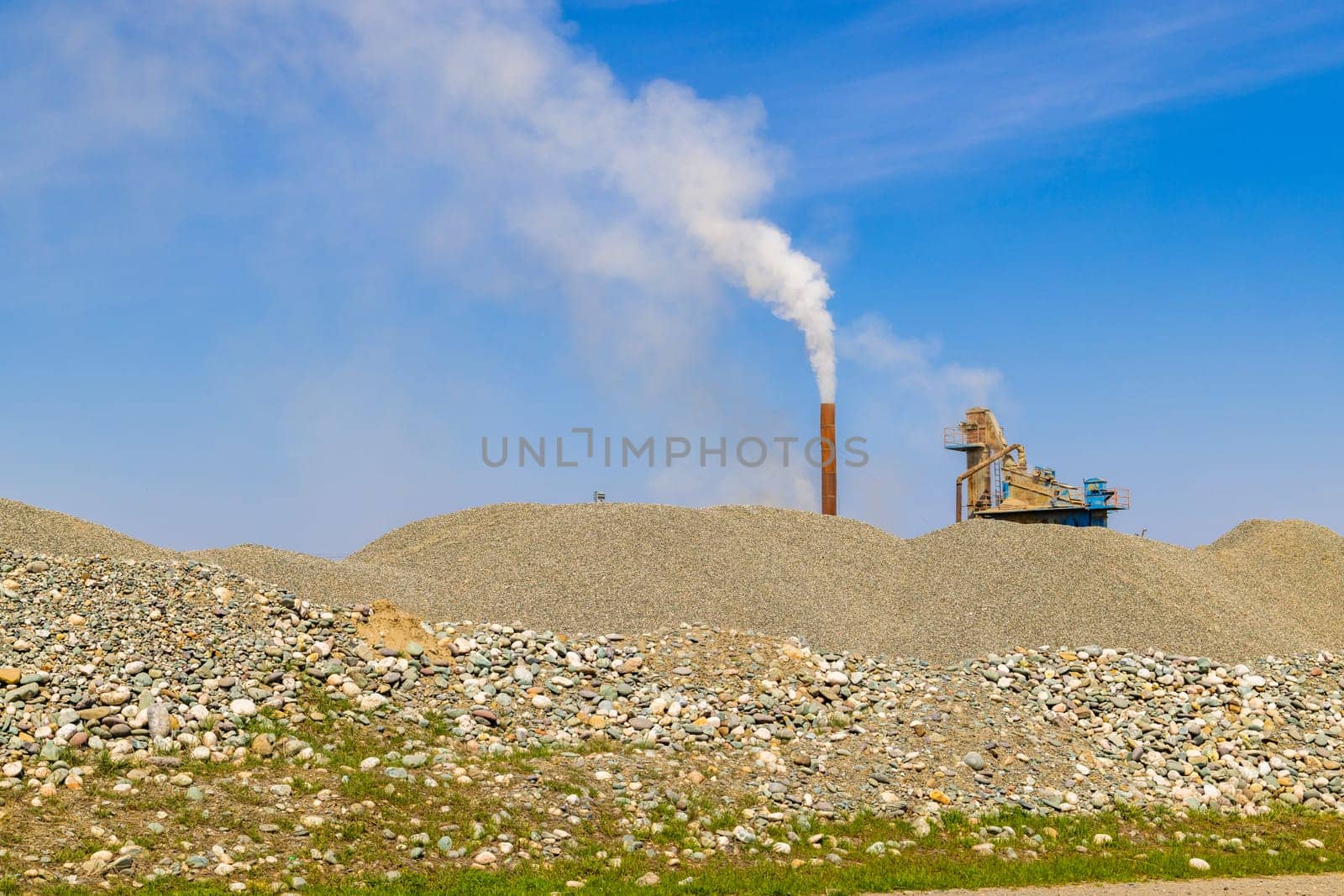 white steam rising up from pipe of small asphalt factory at day time with piles of gravel in a foreground.