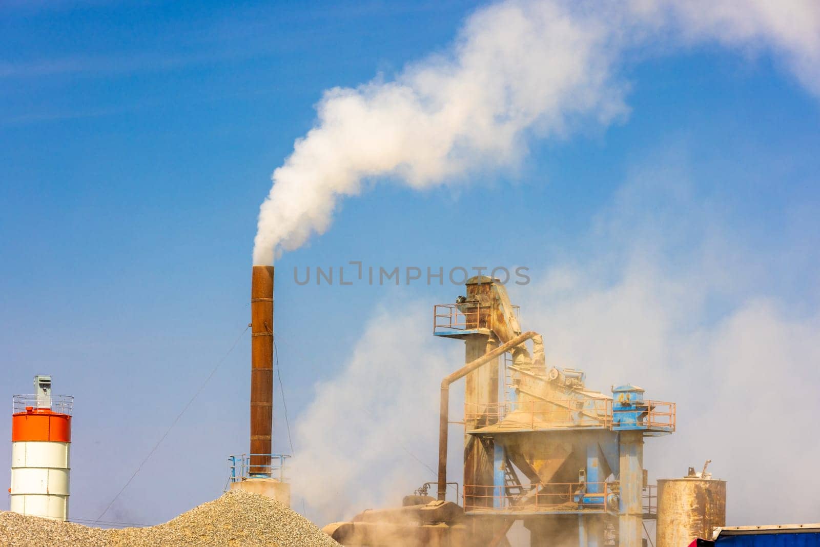 white steam rising up from pipe of small asphalt factory at day time with piles of gravel in a foreground.