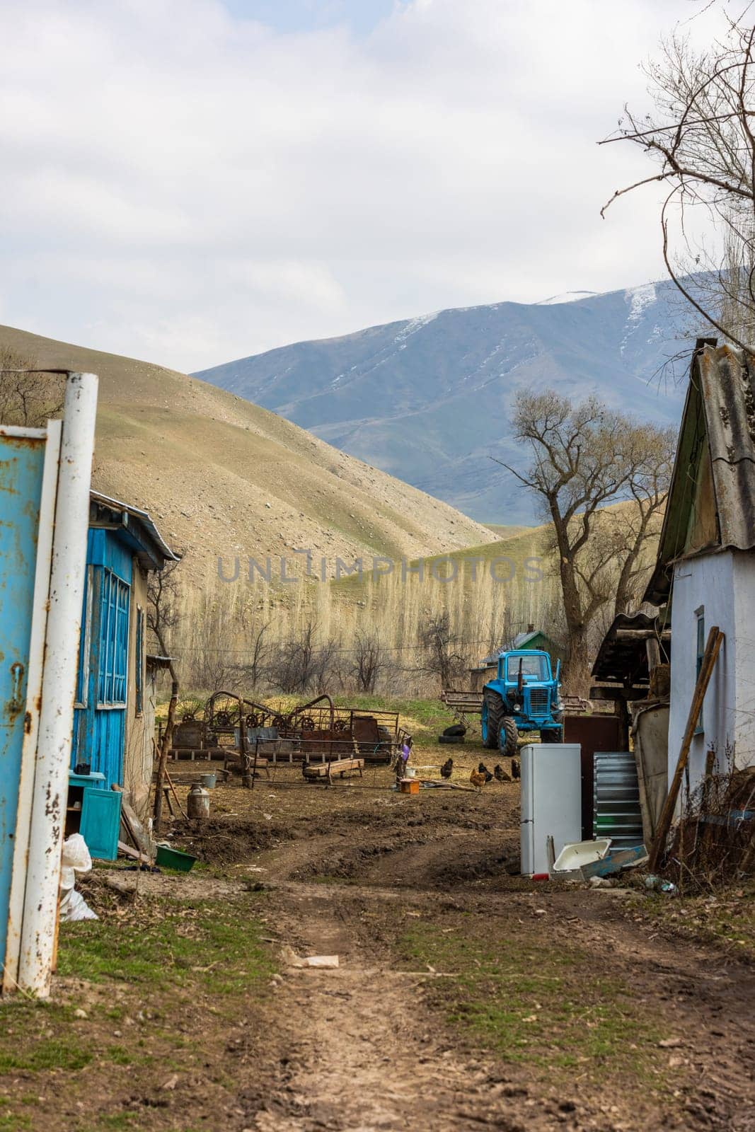 Poor rustic backyard with old blue tractor with dirt road between houses behind rusted steel gate at sunny spring day