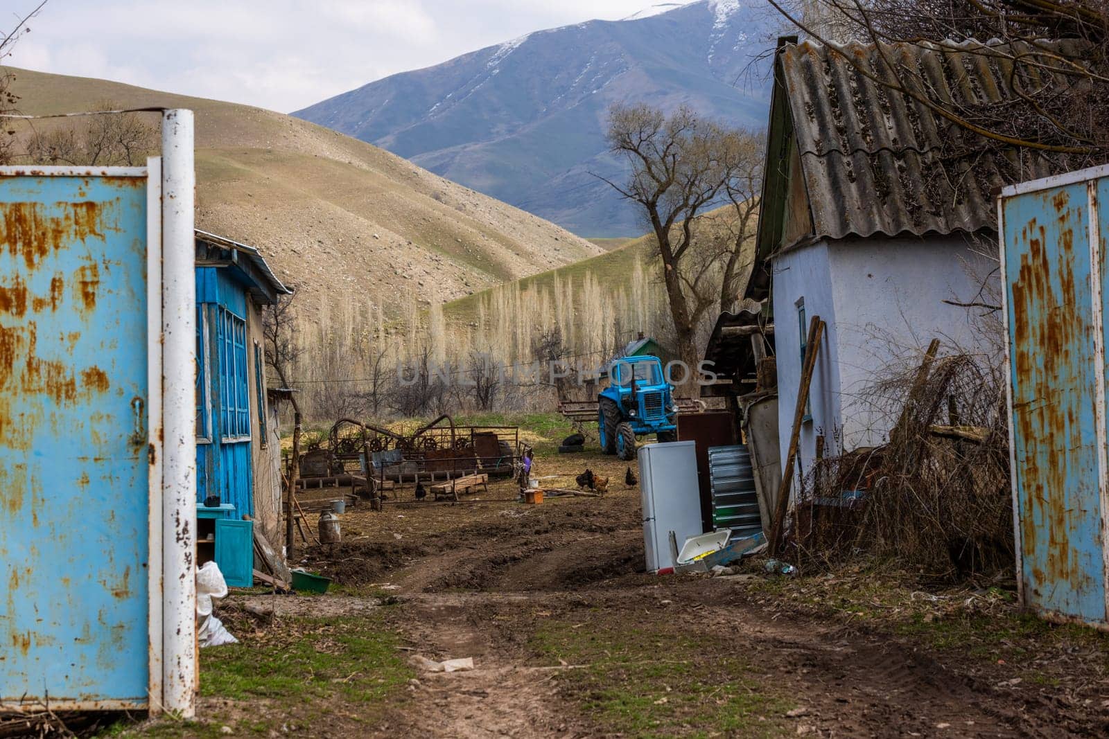 Poor rustic backyard with old blue tractor with dirt road between houses behind rusted steel gate at sunny spring day