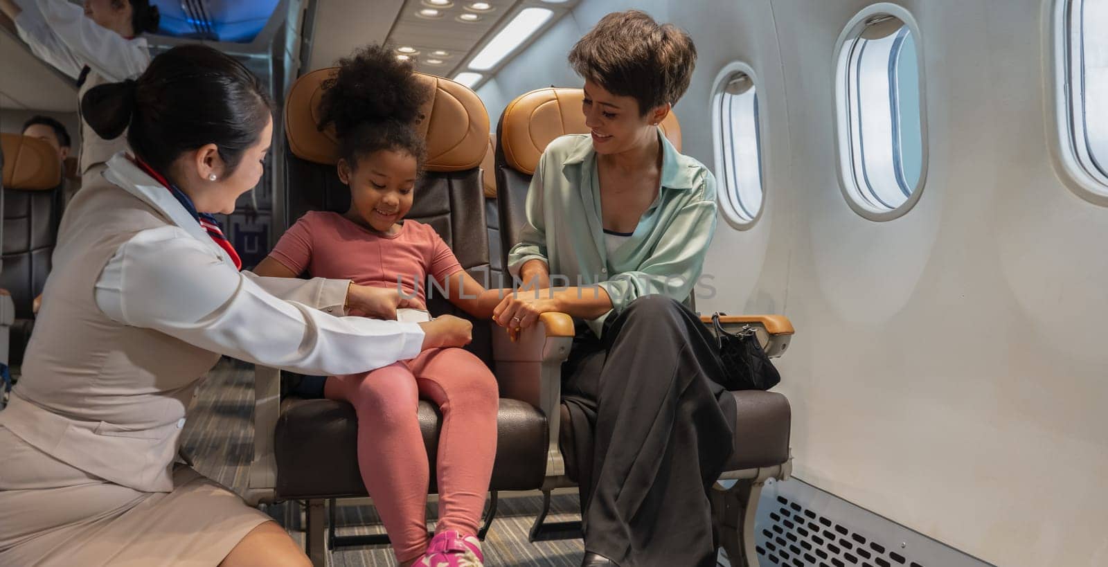 Air hostess helping a kid to fasten her seat belt. Happy family with little girl sitting at airplane. Airline transportation and travel concept.