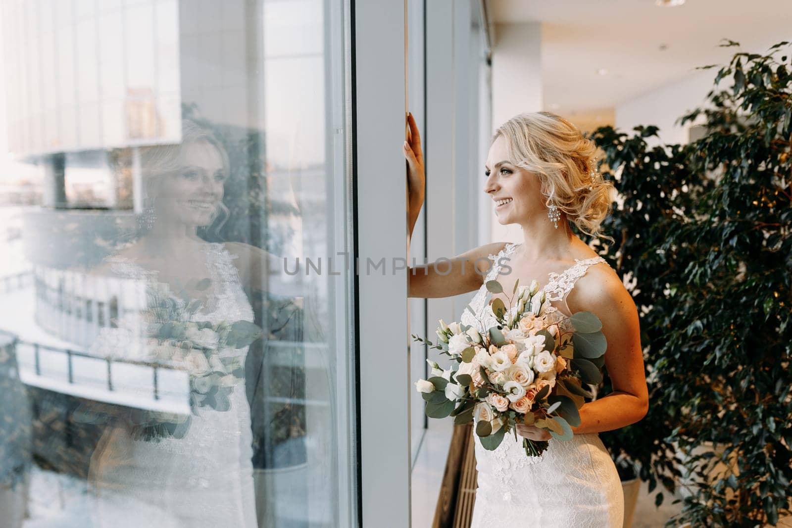 The bride, dressed in a white dress, stands at the window, holding a beautiful bouquet in her hands