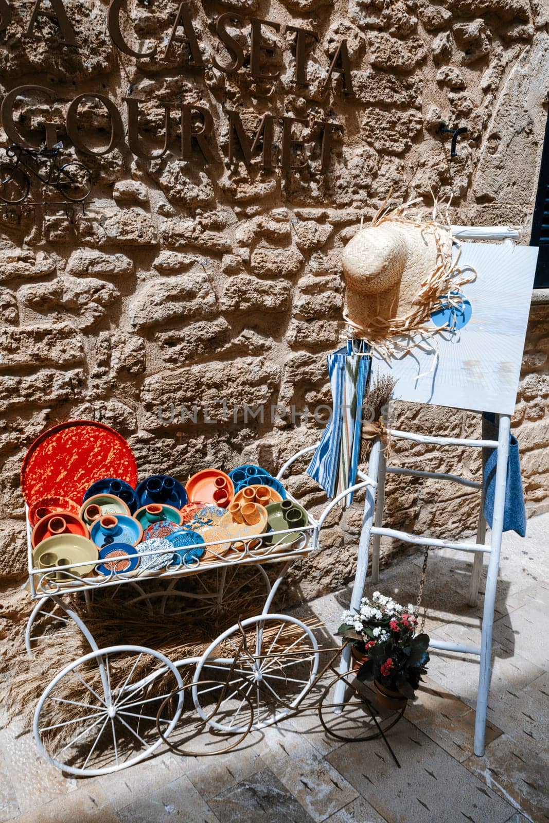 Alcudia Mallorca, Rustic Mediterranean decor: Colorful ceramic plates in vintage metal cart, straw hat on ladder, stone wall background. Traditional European street decoration