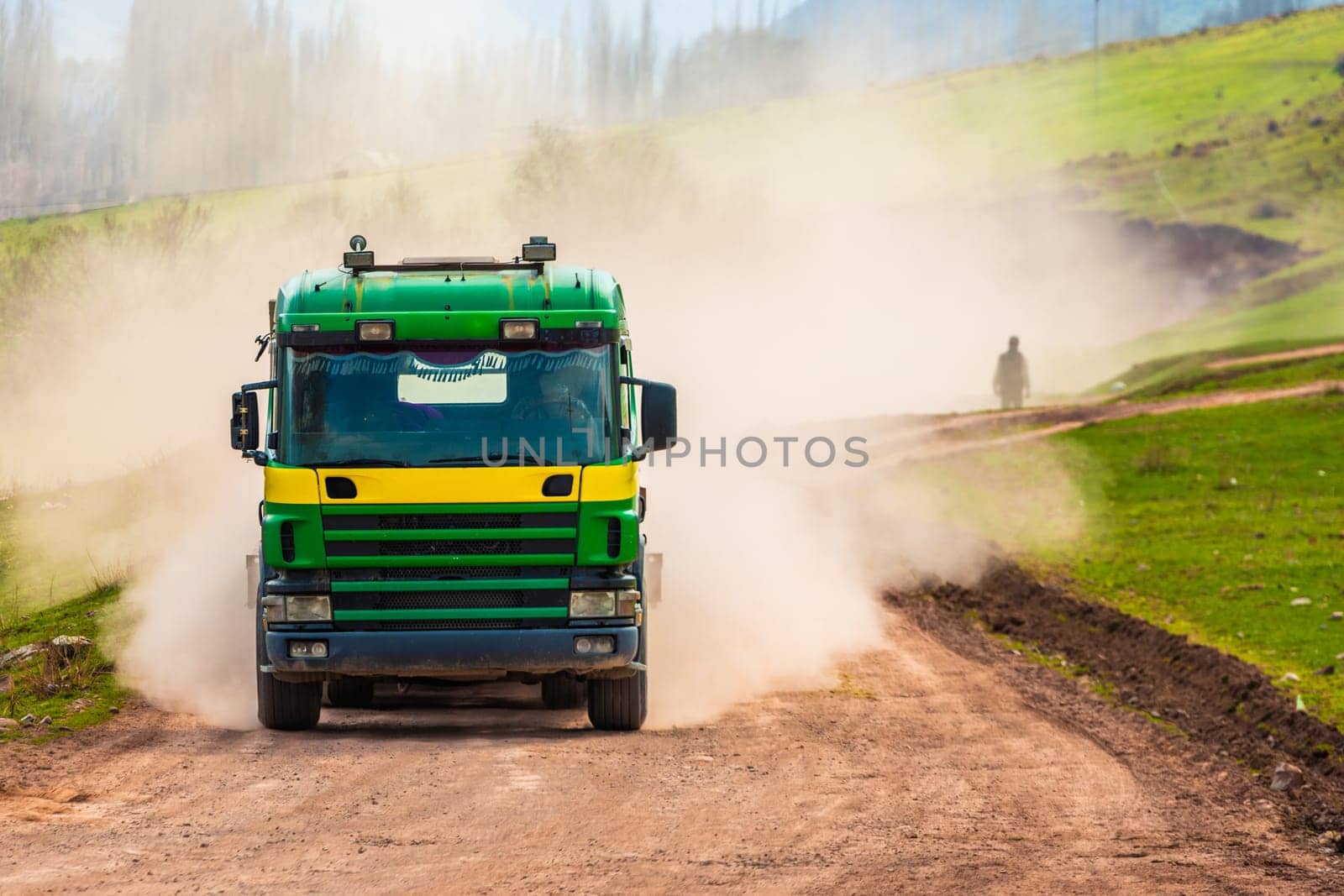 A truck kicks up dust on a dirt road with a pedestrian walking through the haze on a sunny spring day.
