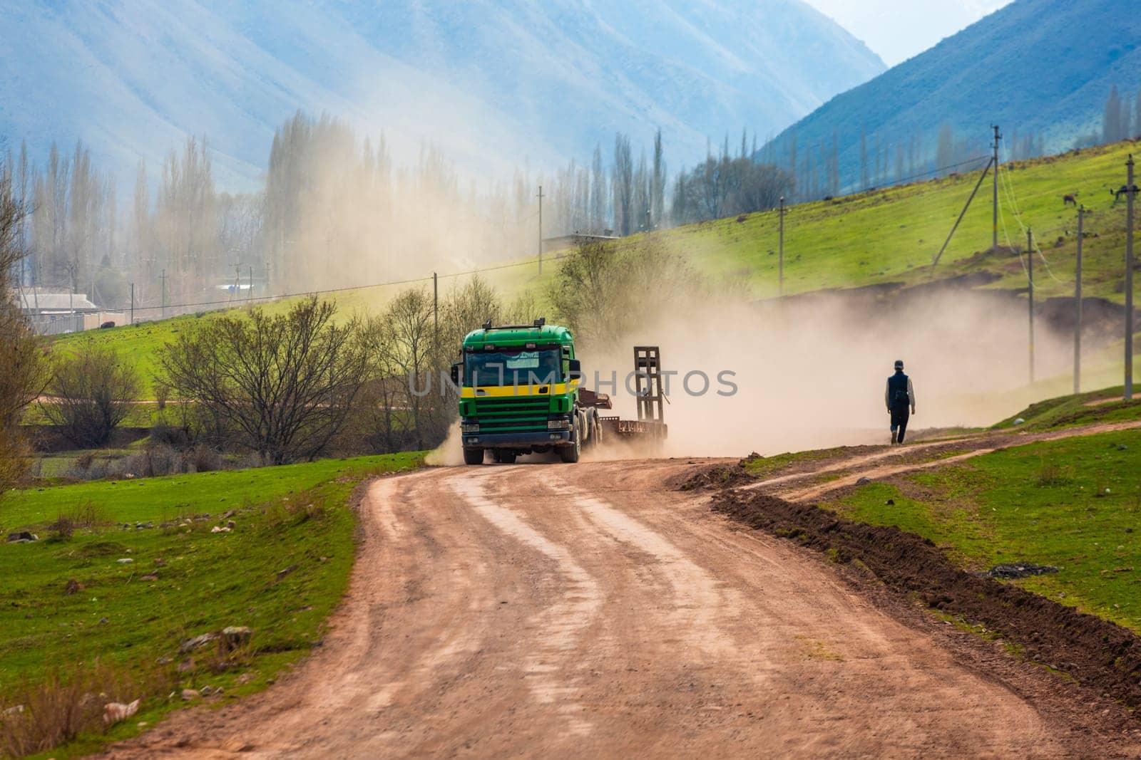 A truck kicks up dust on a dirt road with a pedestrian walking through the haze on a sunny spring day.