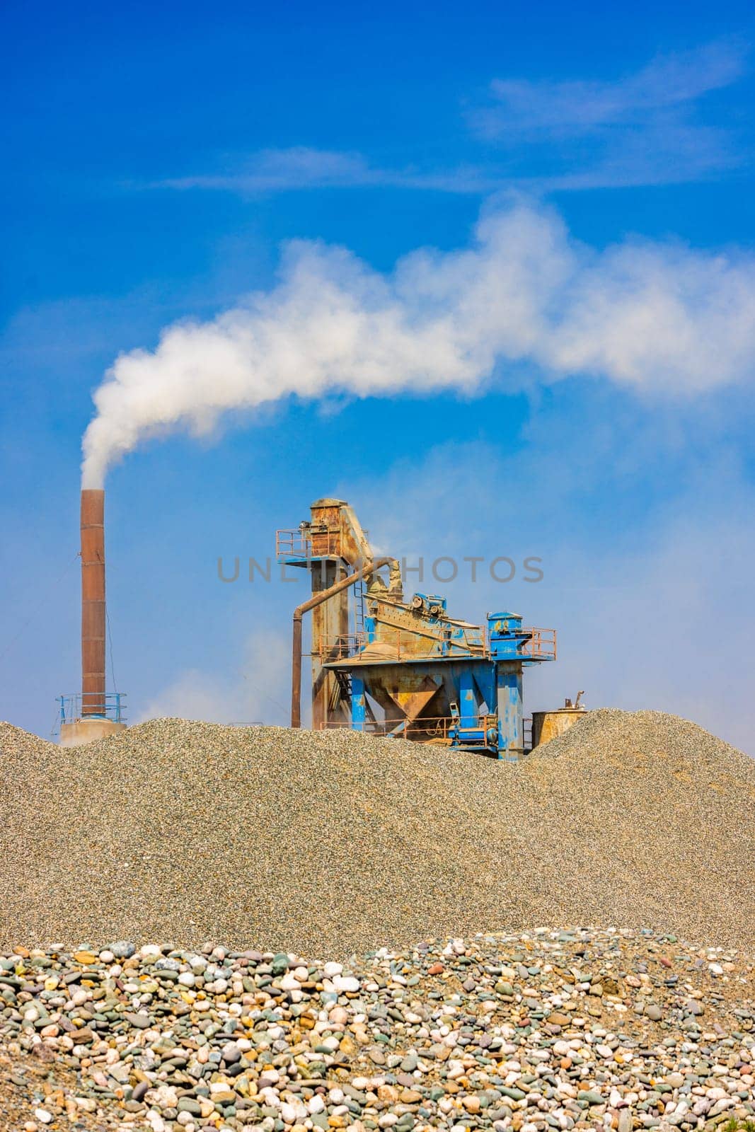 white steam rising up from pipe of small asphalt factory at day time with piles of gravel in a foreground.
