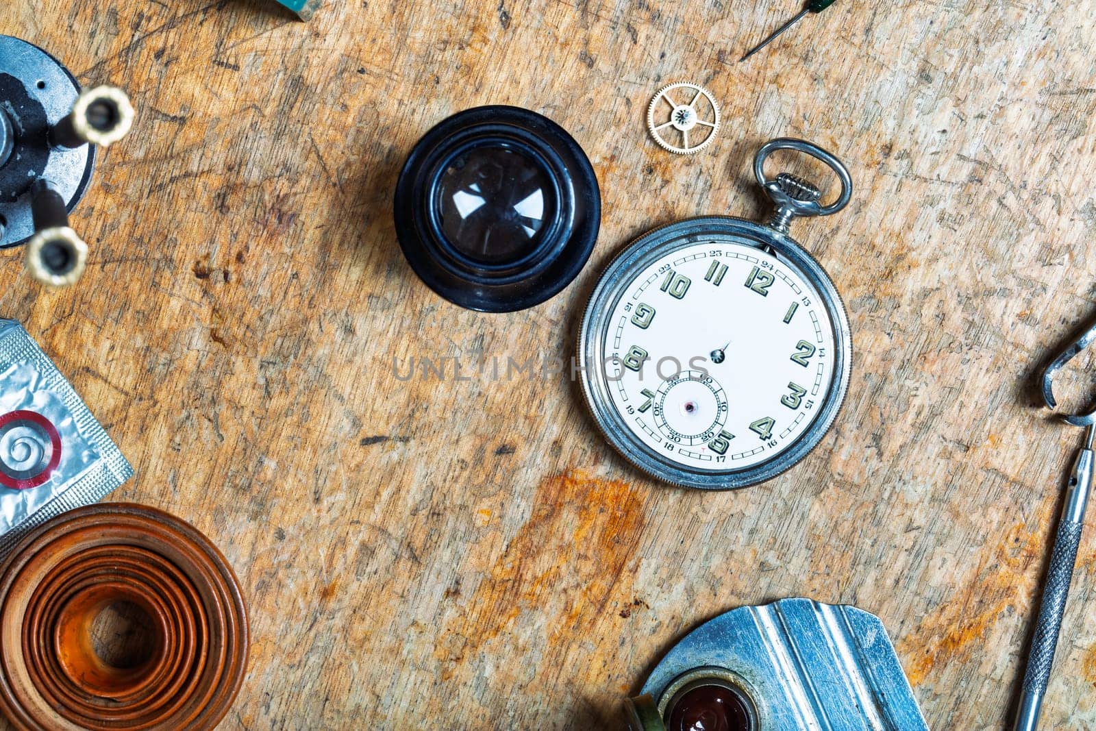 assortment of watchmaking tools surrounds a pocket watch on a worn wooden surface, displaying components like a gasket coil, magnifier, and gear, highlighting precision work