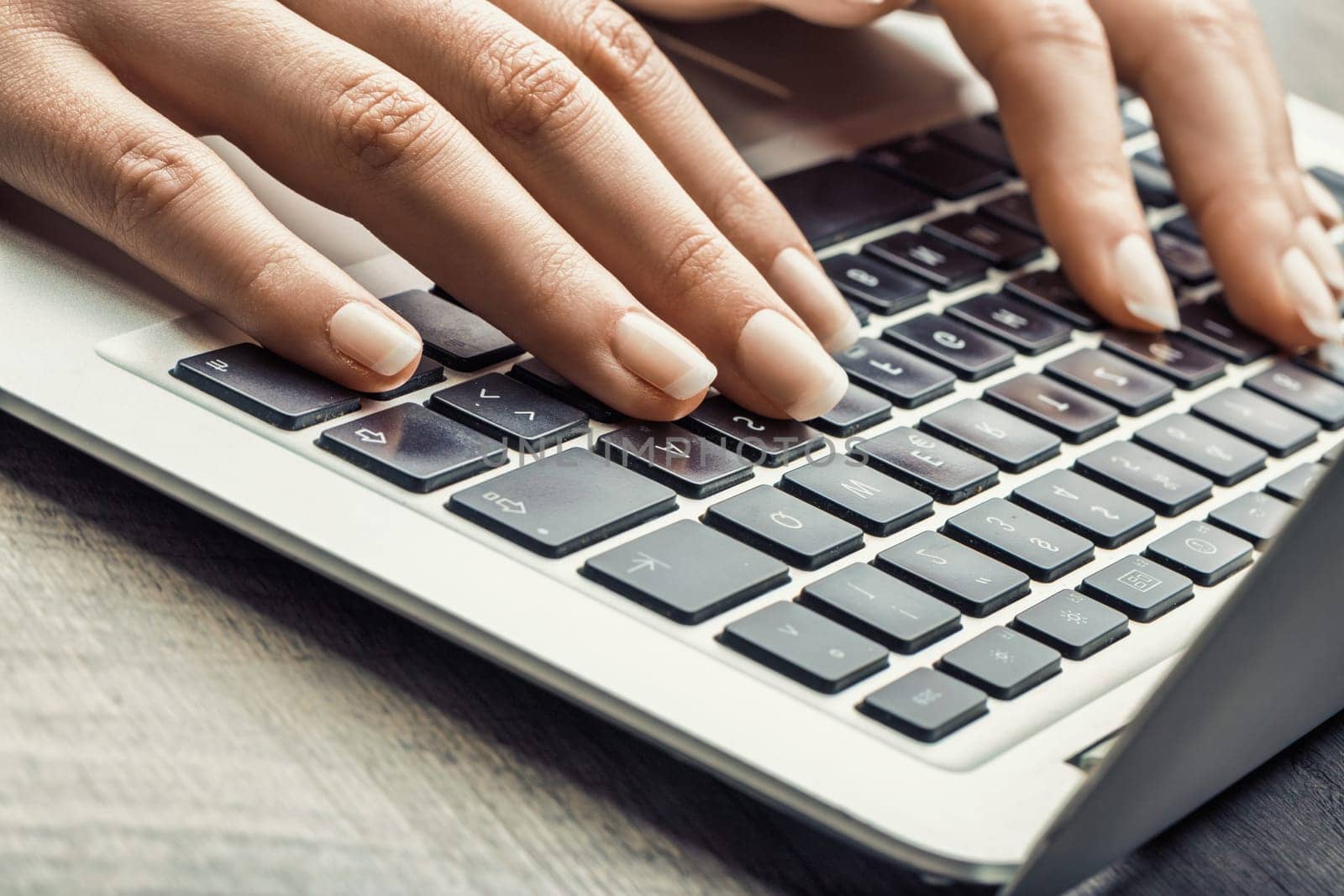 Hand with light skin and polished nails typing on a laptop, with a soft-toned background and coffee cup, adding focus to a peaceful workspace