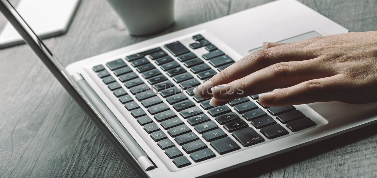 Light skin and well-manicured nails on a hand pressing a laptop key. The background shows a coffee cup, adding to the focused, calm workspace ambiance